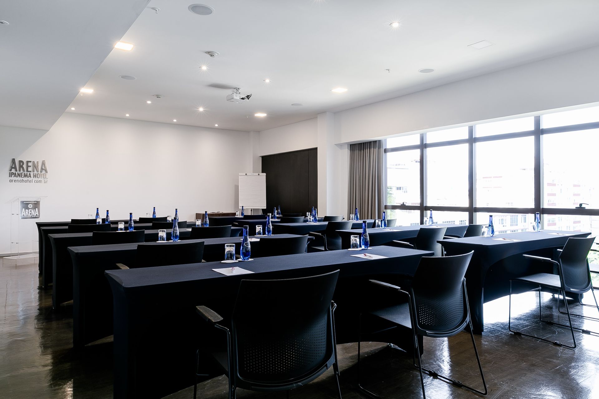 Sala de conferências moderna e vazia, com mesas e cadeiras pretas, janelas amplas e iluminação embutida no teto.