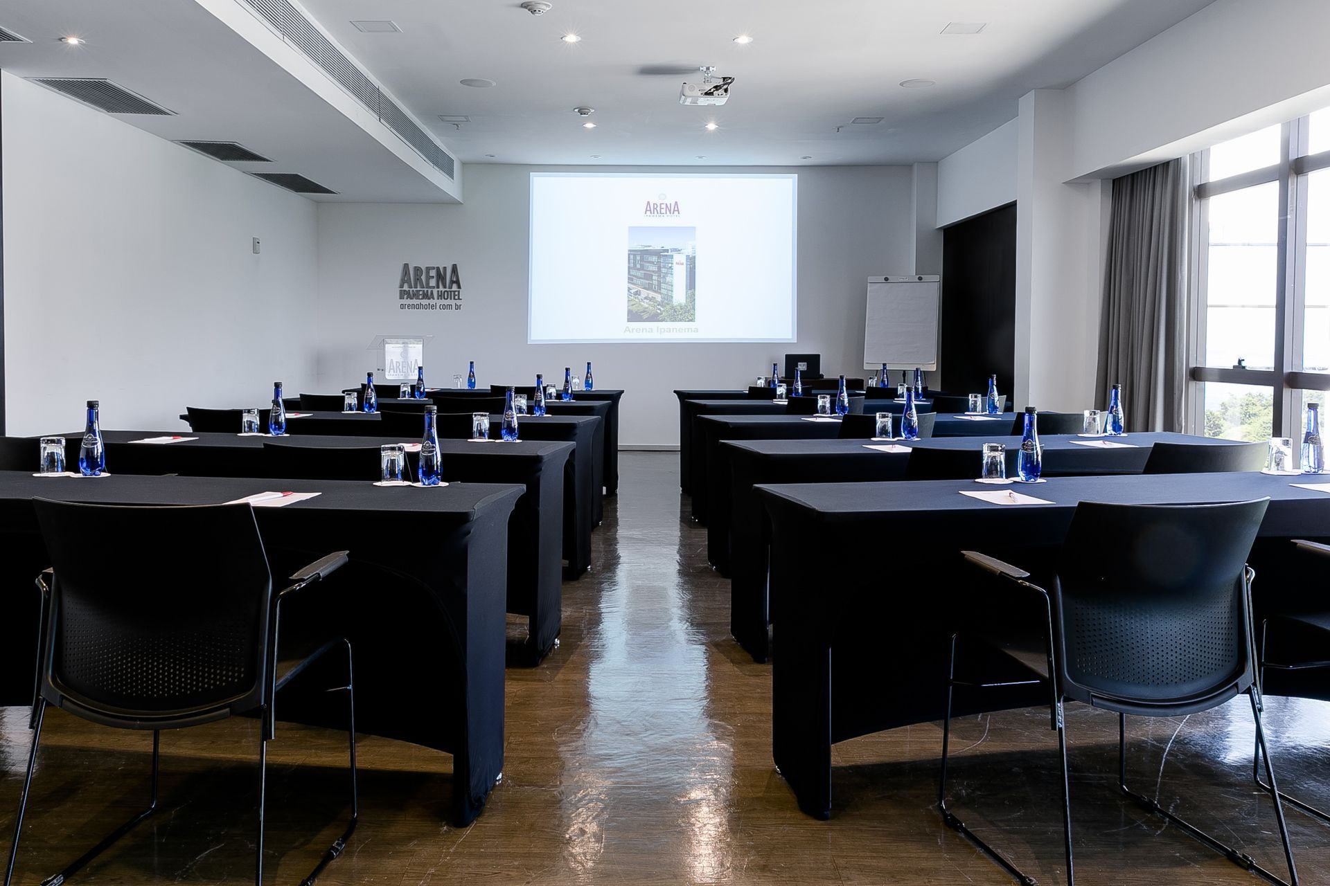 Sala de treinamento vazia com fileiras de mesas e cadeiras pretas voltadas para uma tela de projeção, iluminada pela luz do dia.