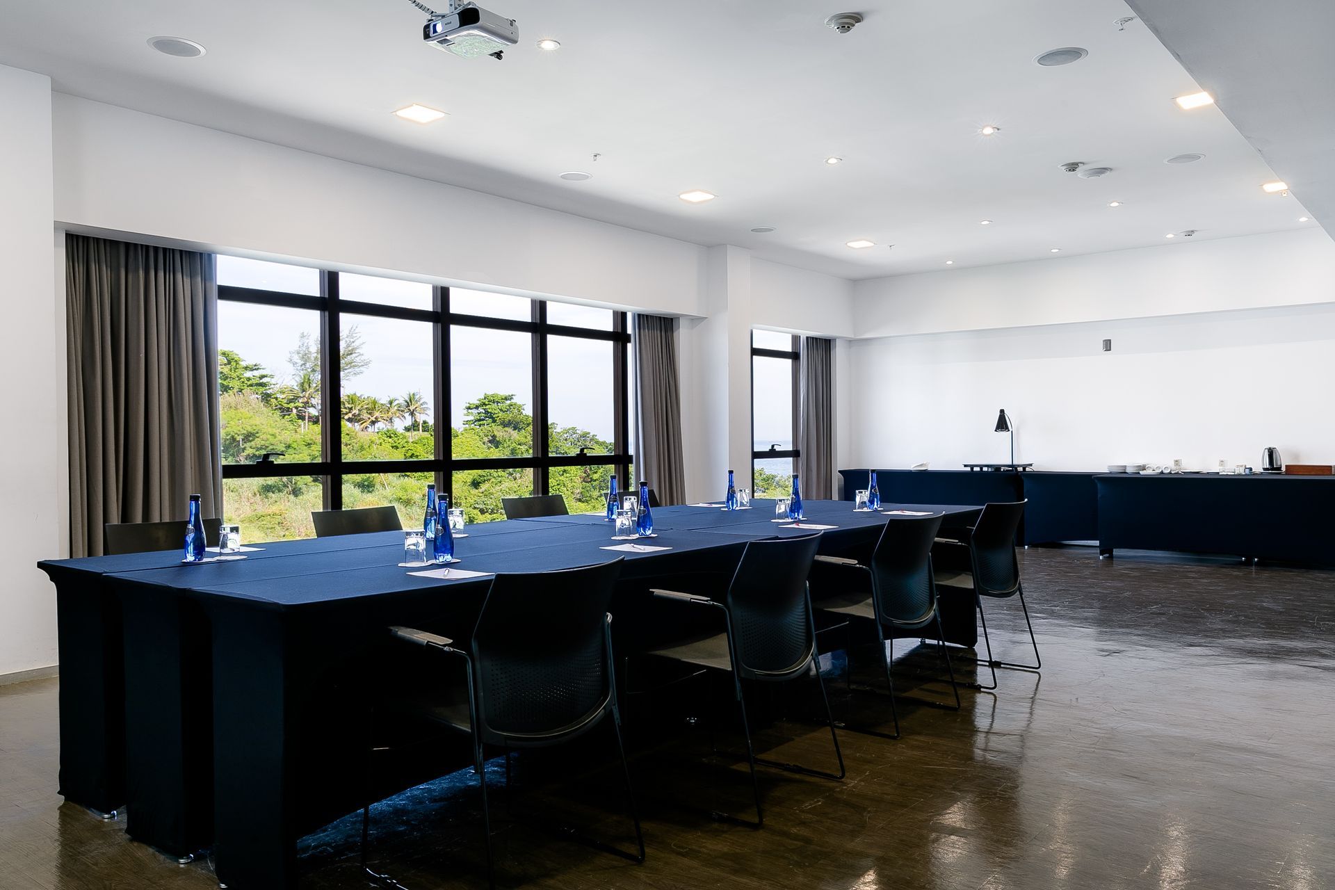 Sala de conferências moderna e vazia, com mesas compridas, cadeiras pretas e grandes janelas com vista para a vegetação.