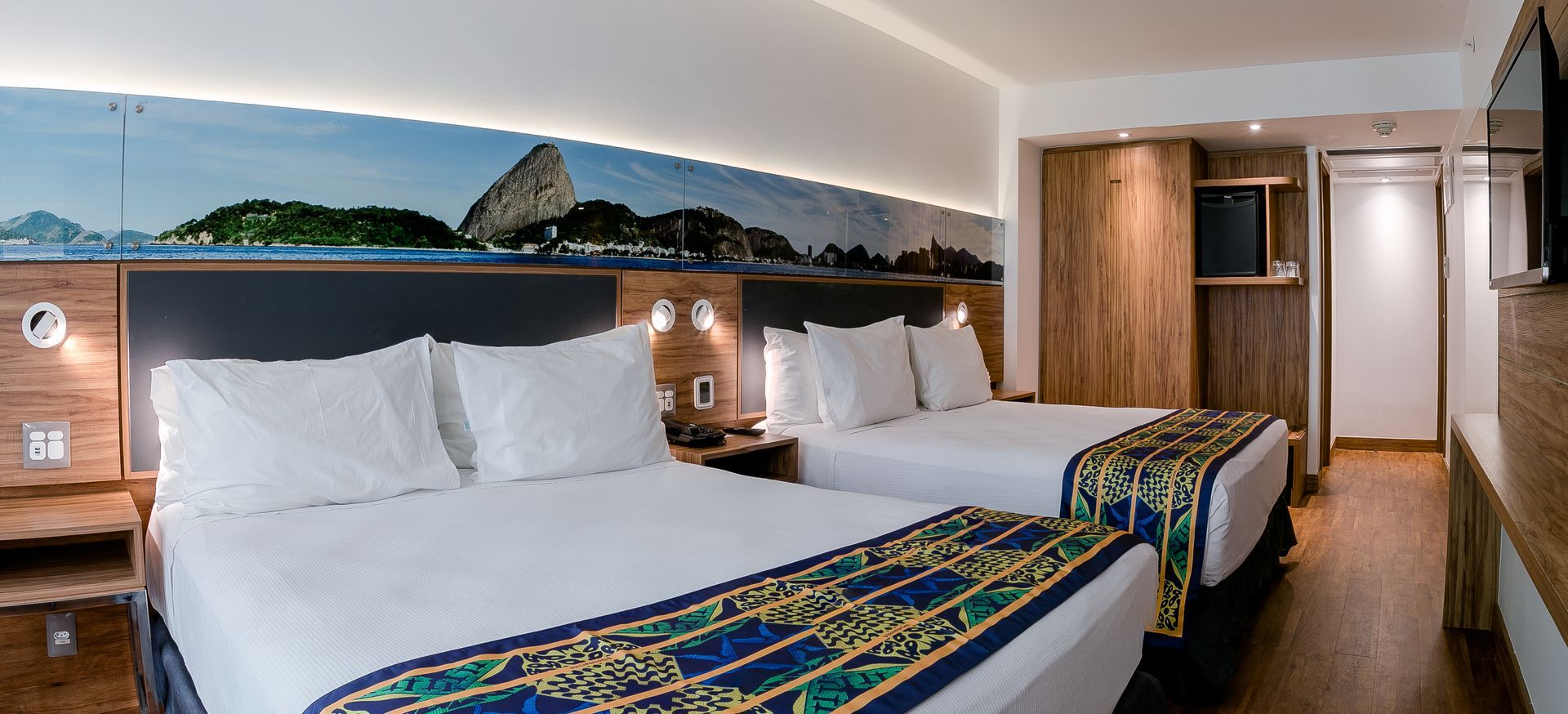 Quarto de hotel com duas camas bem arrumadas, piso de madeira e um mural de montanha acima das cabeceiras.