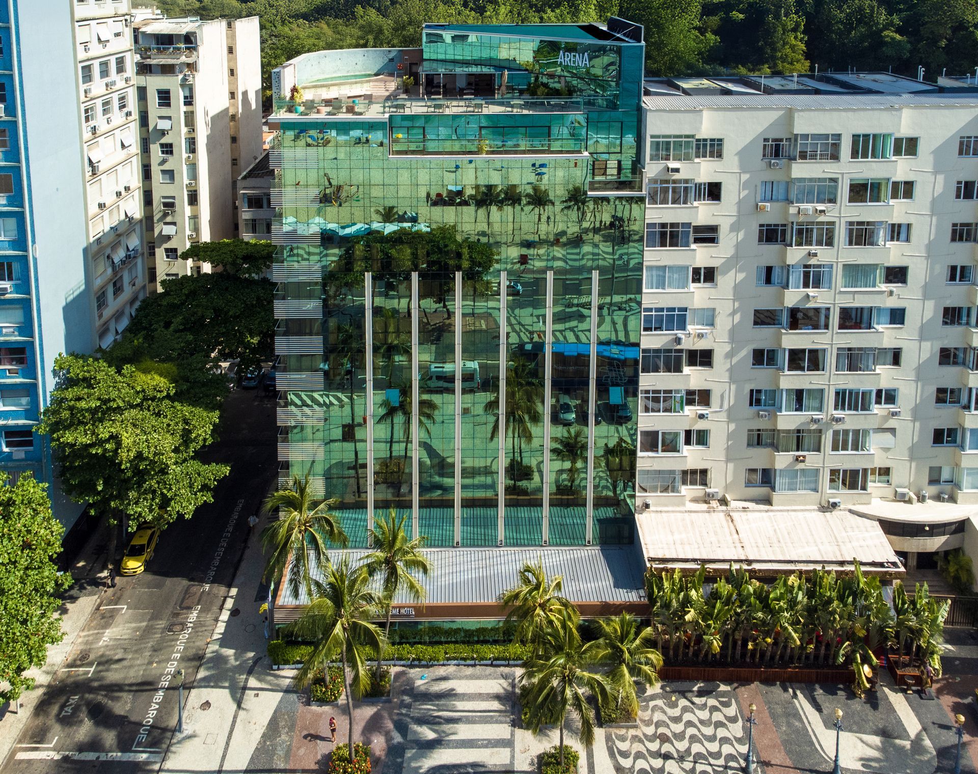 Prédio moderno com fachada de vidro e jardim verde na cobertura, entre blocos de apartamentos brancos e palmeiras.