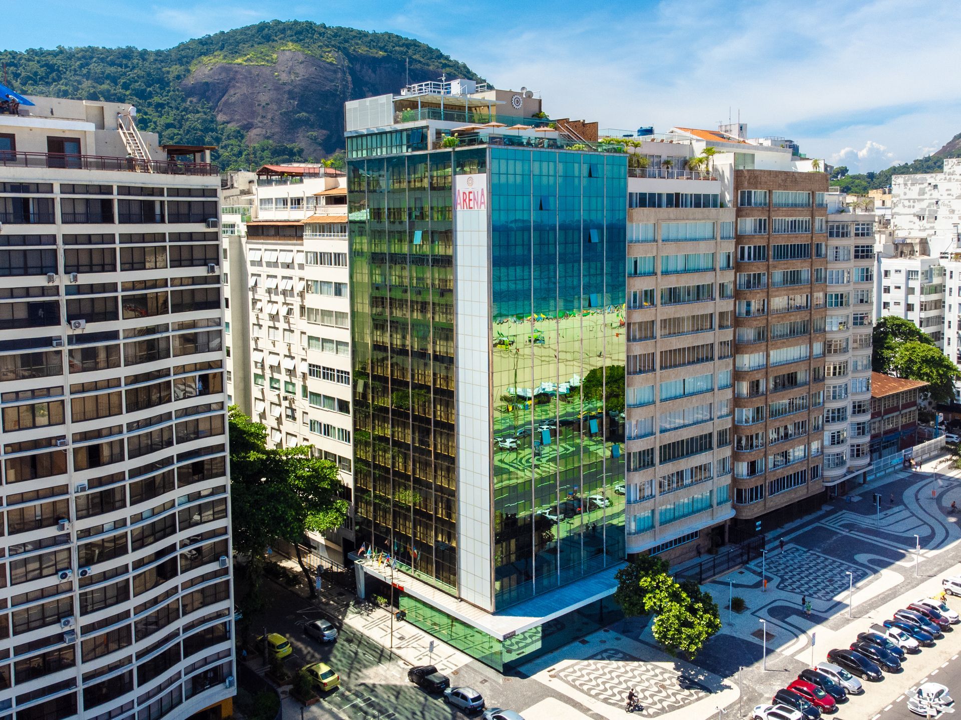 Quarteirão com um prédio de escritórios de vidro verde refletivo ao lado de apartamentos altos e uma rua com carros estacionados.