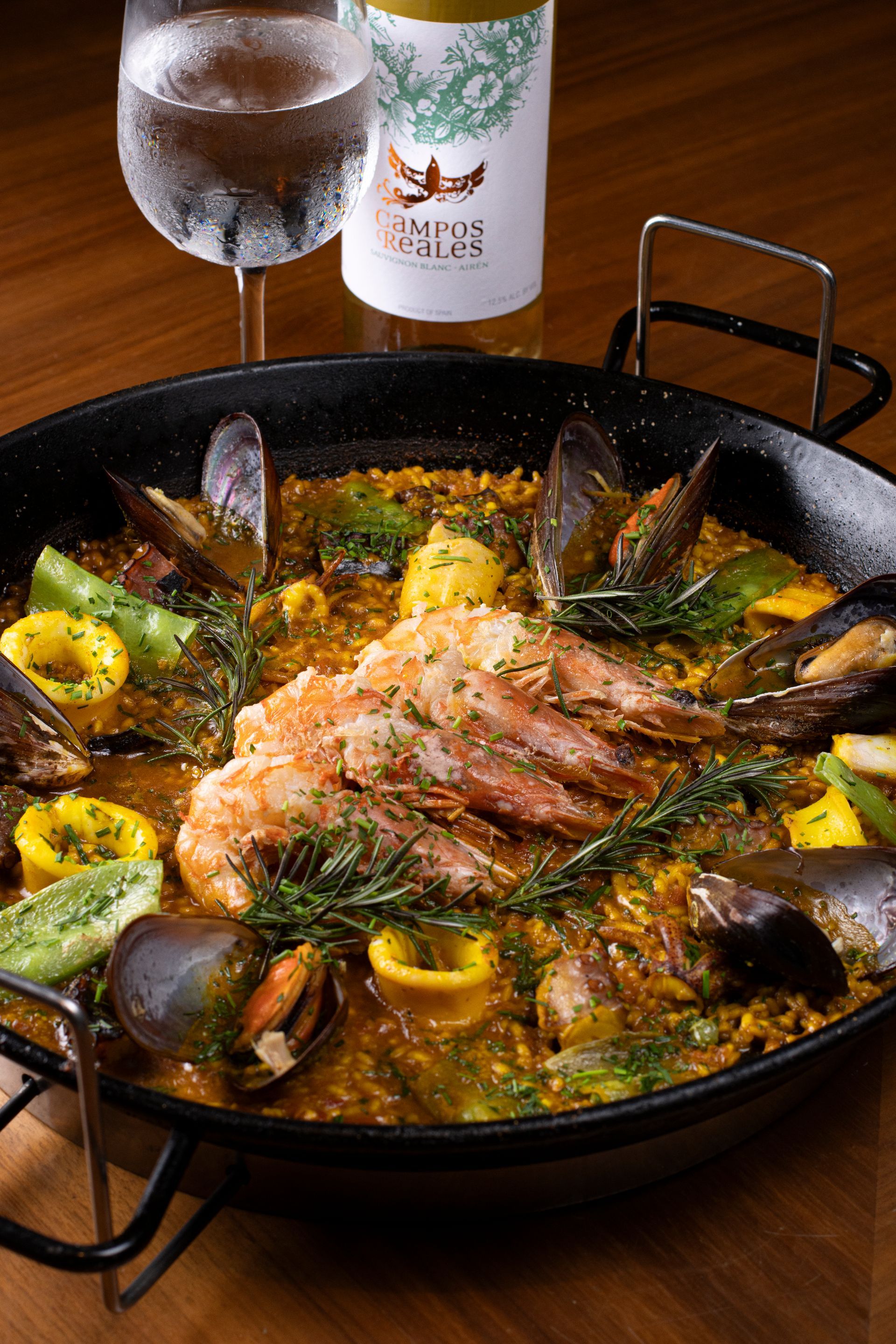 Panela de paella com frutos do mar e fatias de limão sobre a mesa, ao lado de uma taça e uma garrafa de vinho.