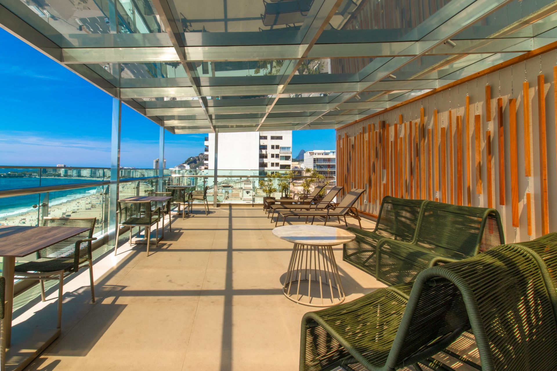 Terraço na cobertura com vista para o mar, cobertura de vidro, cadeiras de vime e mesas ensolaradas perto da orla.