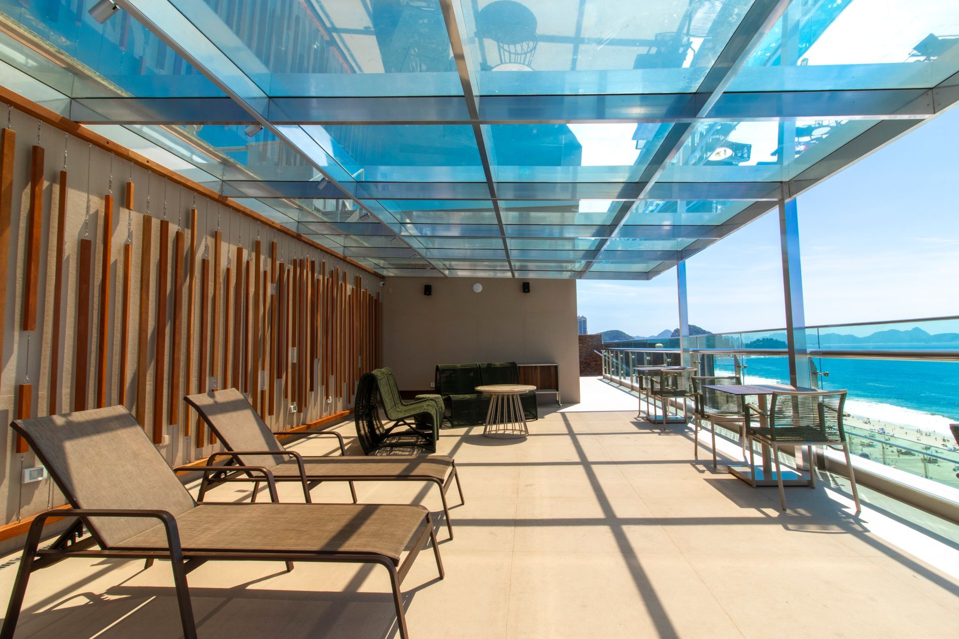 Terraço ensolarado à beira-mar com espreguiçadeiras sob um toldo de vidro azul e vista para o oceano.