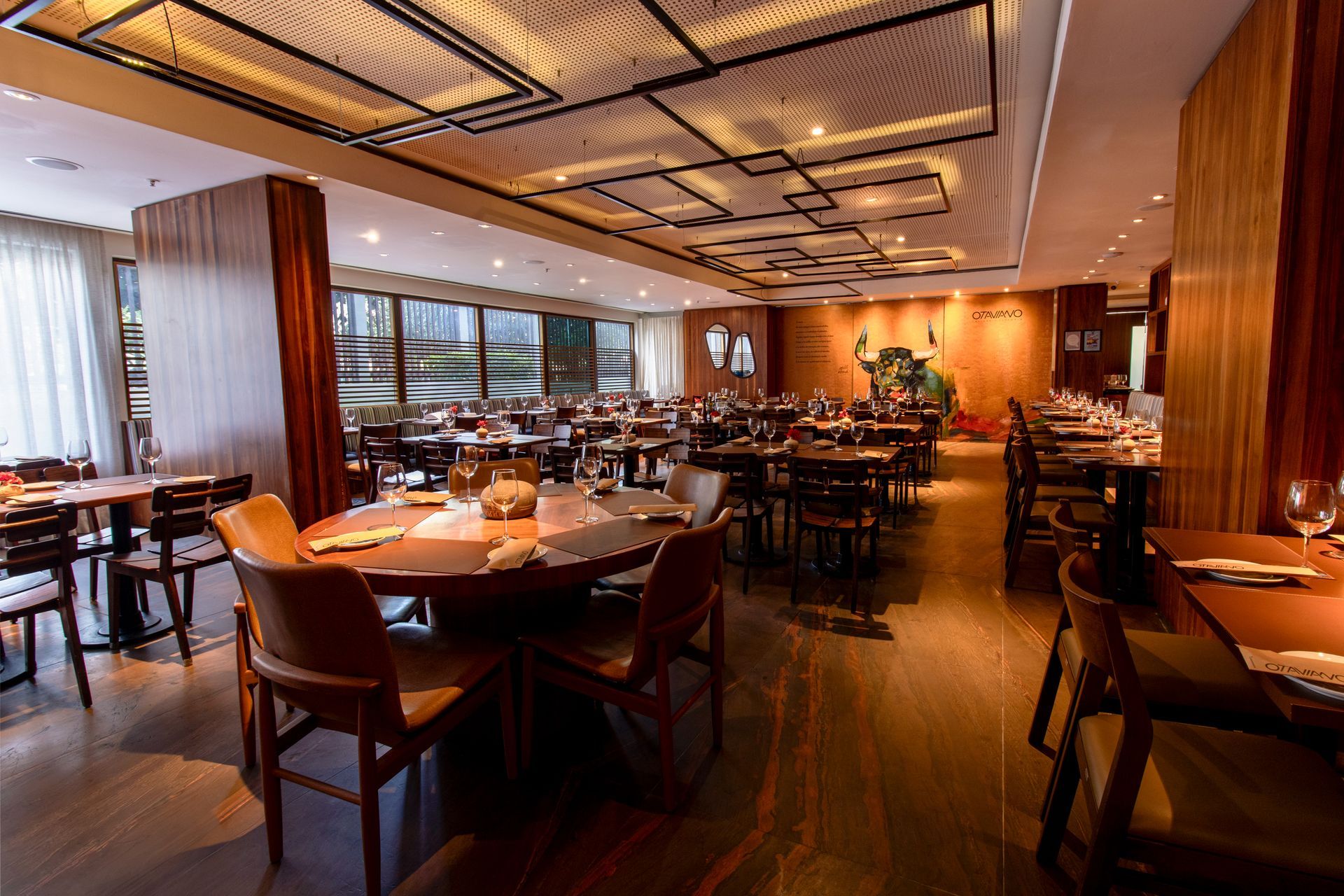 Restaurante moderno com iluminação aconchegante, mesas e cadeiras postas, e grandes janelas ao entardecer.
