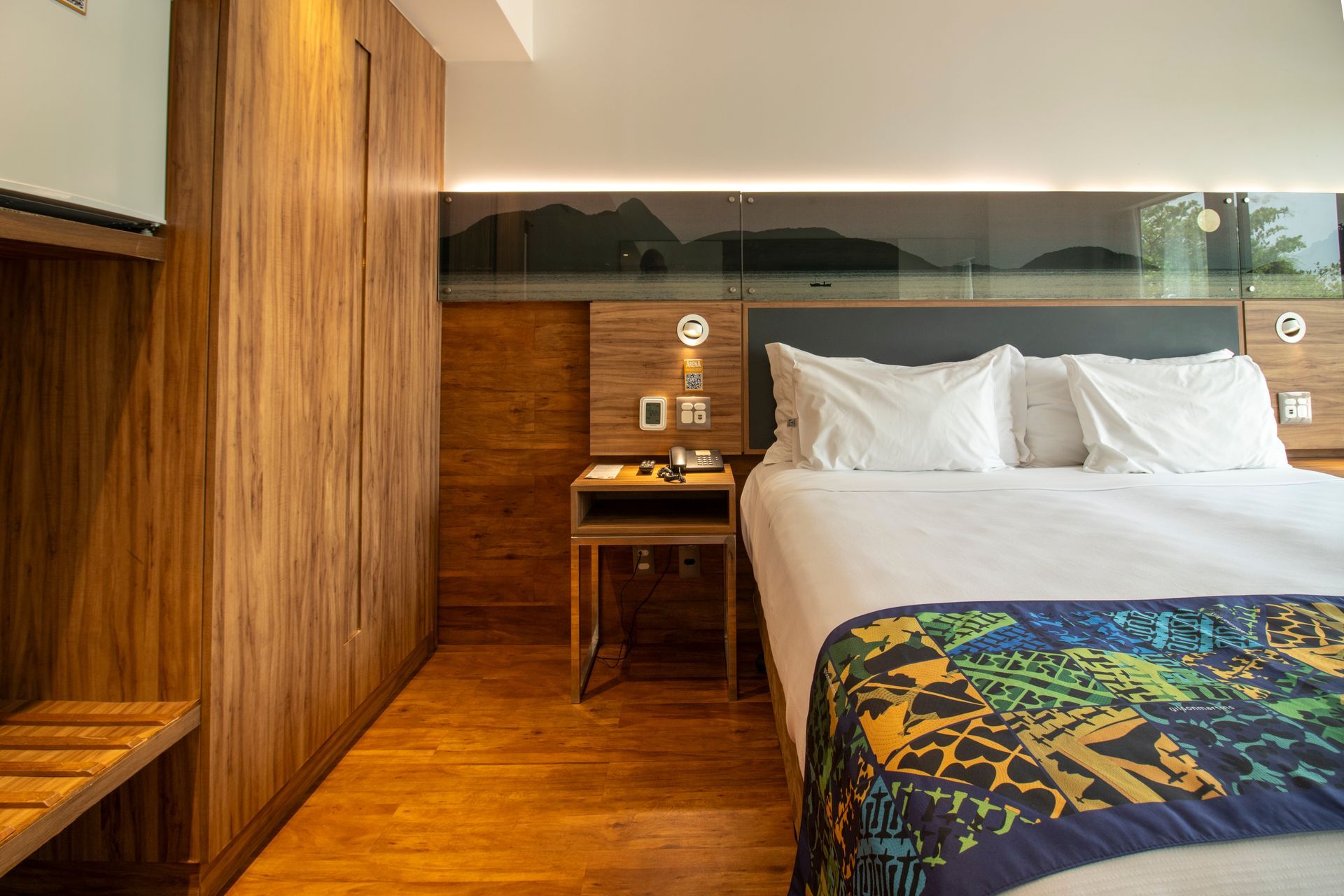 Quarto de hotel moderno com painéis de madeira, cama grande, mesa de cabeceira e cobertor colorido.