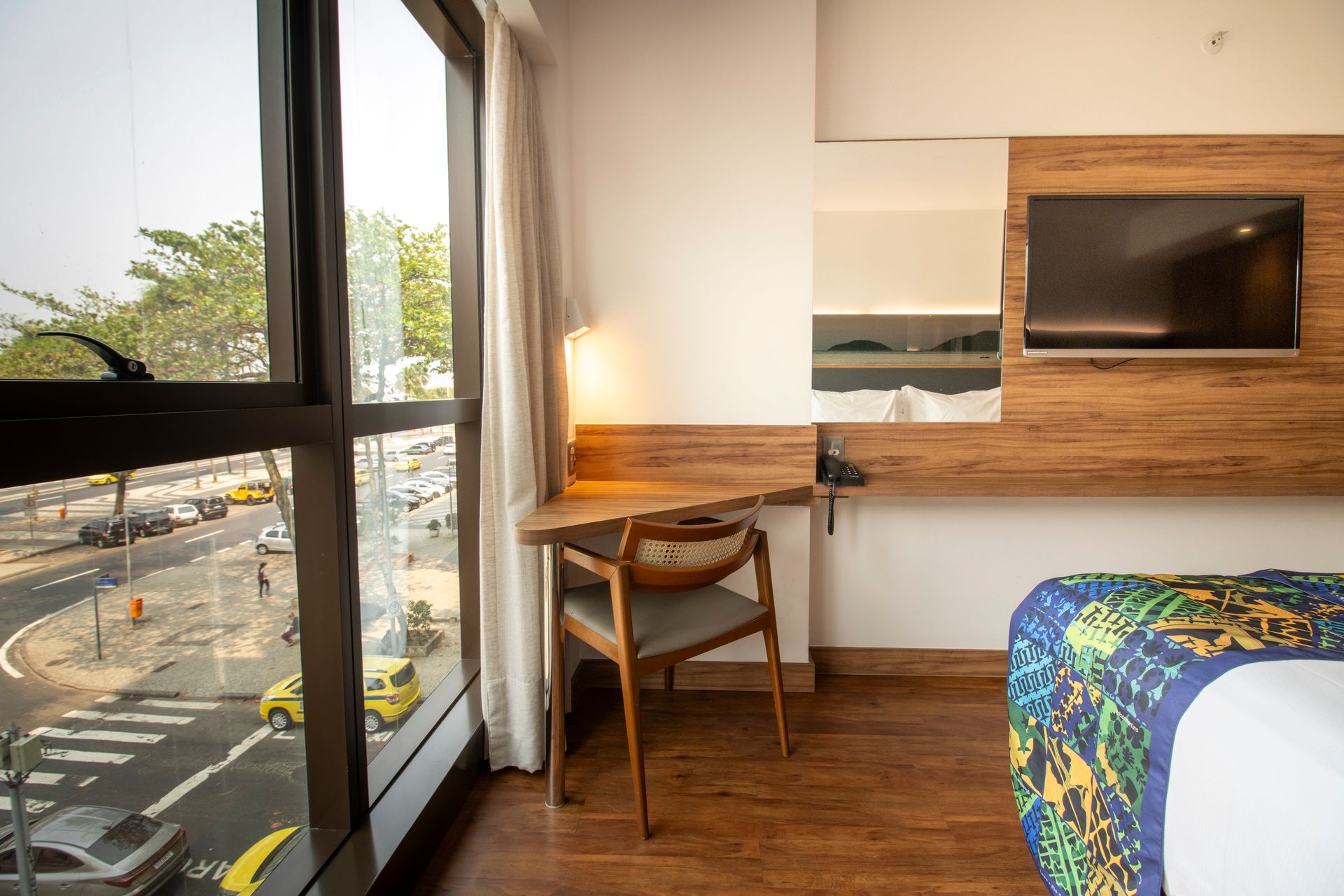 Quarto de hotel com escrivaninha junto a uma grande janela, piso de madeira e uma cama à direita.