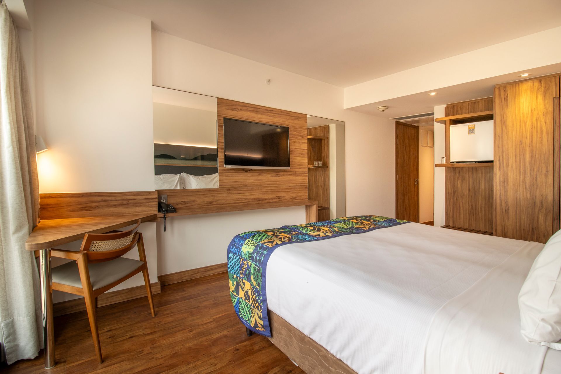 Quarto de hotel moderno com cama, escrivaninha, TV de parede e detalhes em madeira aconchegantes.