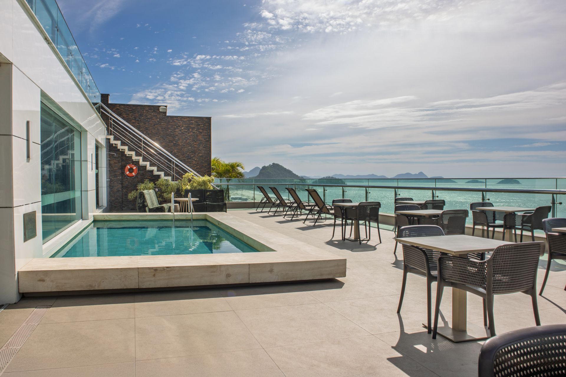Terraço com piscina na cobertura, espreguiçadeiras, mesas e vista para o mar sob um céu claro.