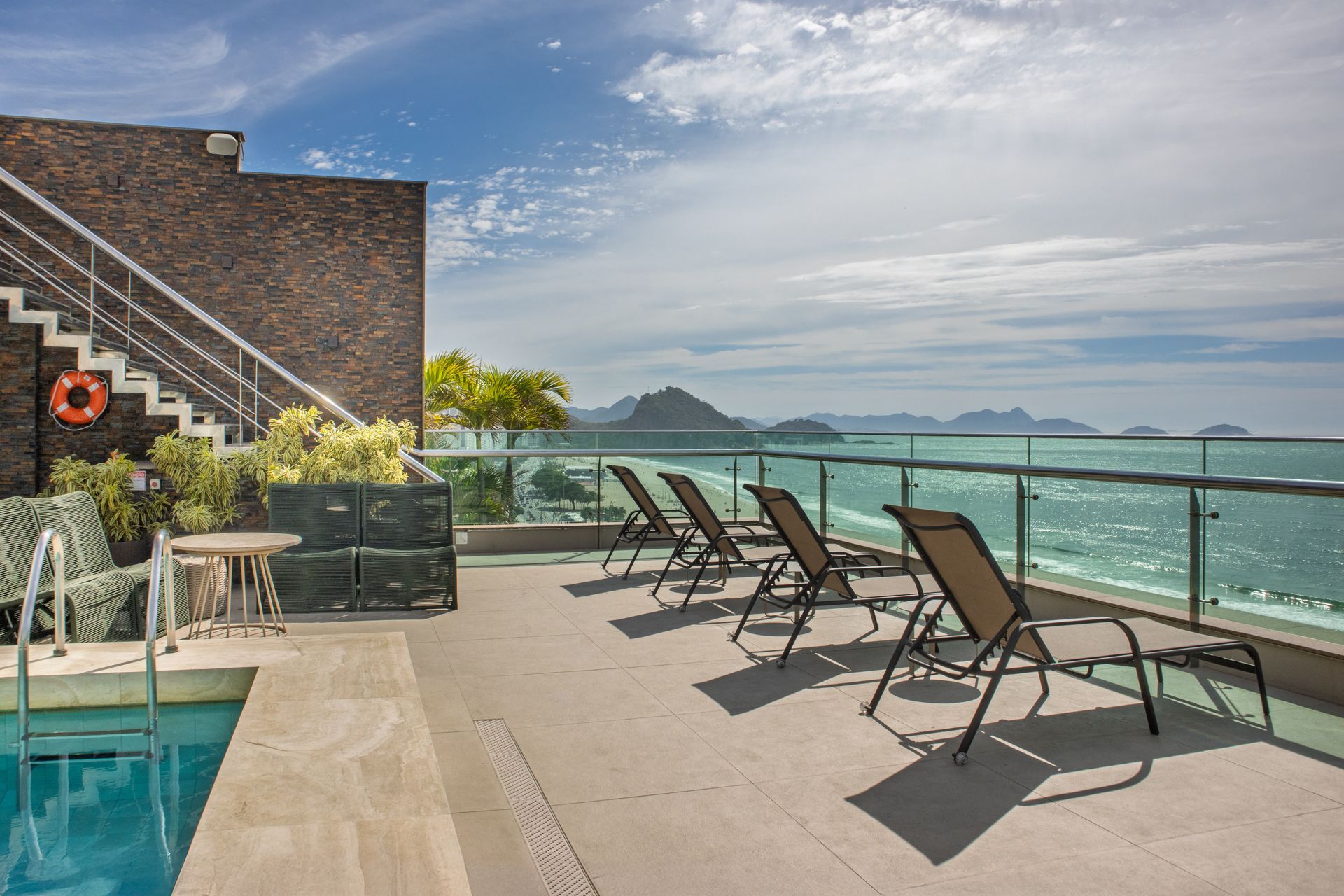 Terraço à beira da piscina com espreguiçadeiras com vista para a água turquesa e montanhas distantes sob um céu azul.