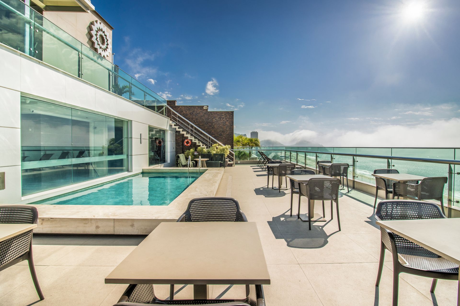 Terraço ensolarado na cobertura com piscina, mesas, cadeiras, guarda-corpo de vidro e vista para o mar.