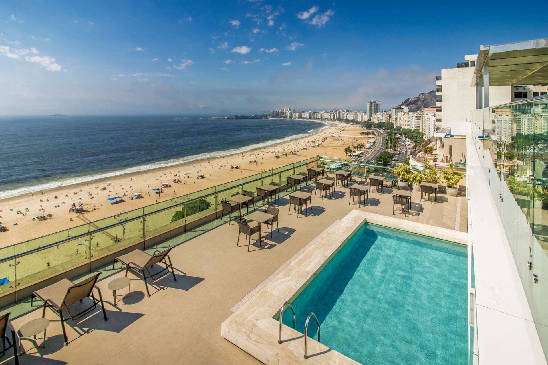 Terraço de resort à beira-mar com piscina, espreguiçadeiras e praia de areia sob um céu azul límpido.