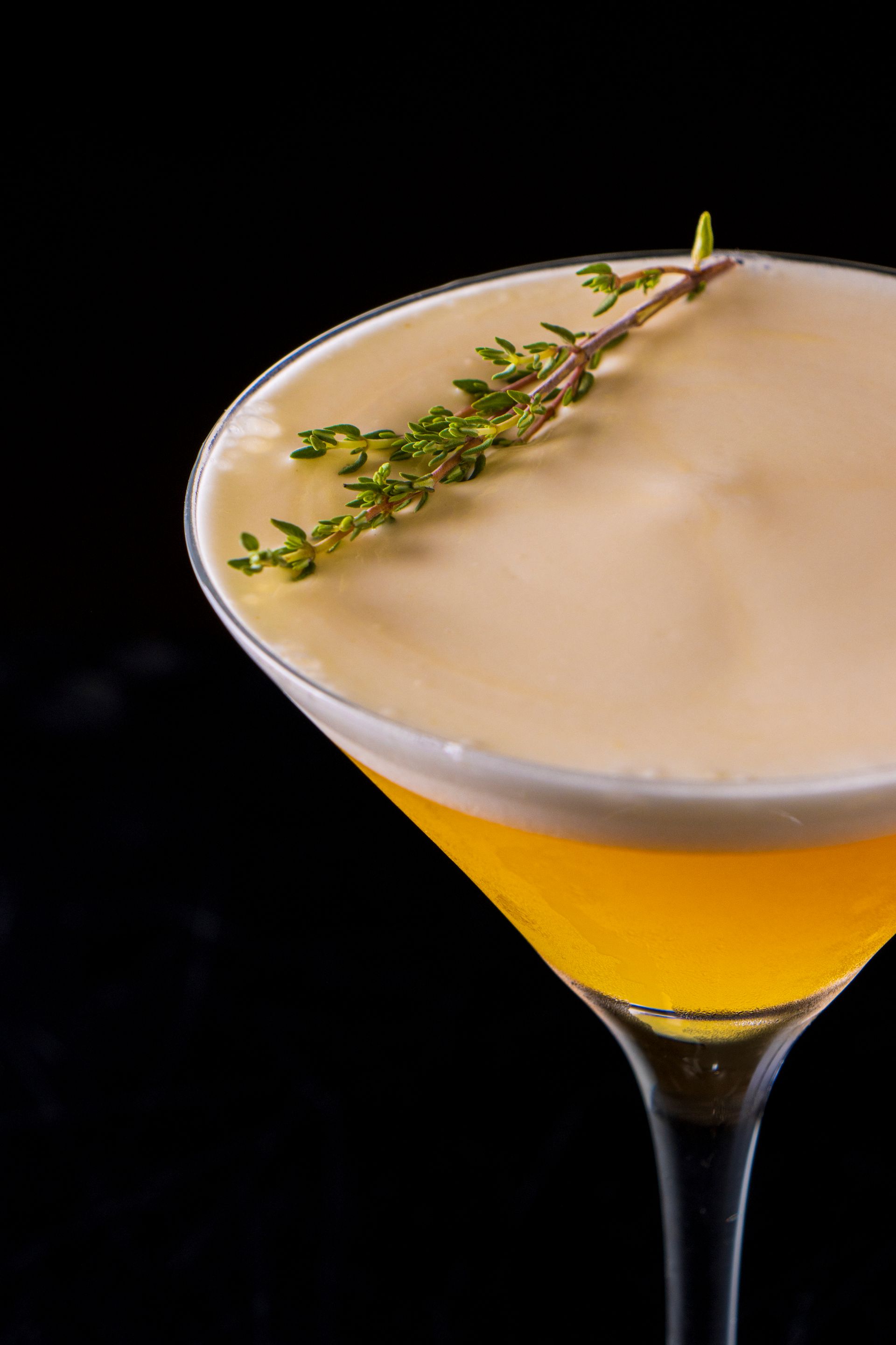 Coquetel amarelo em uma taça de martini, decorado com tomilho sobre um fundo preto.