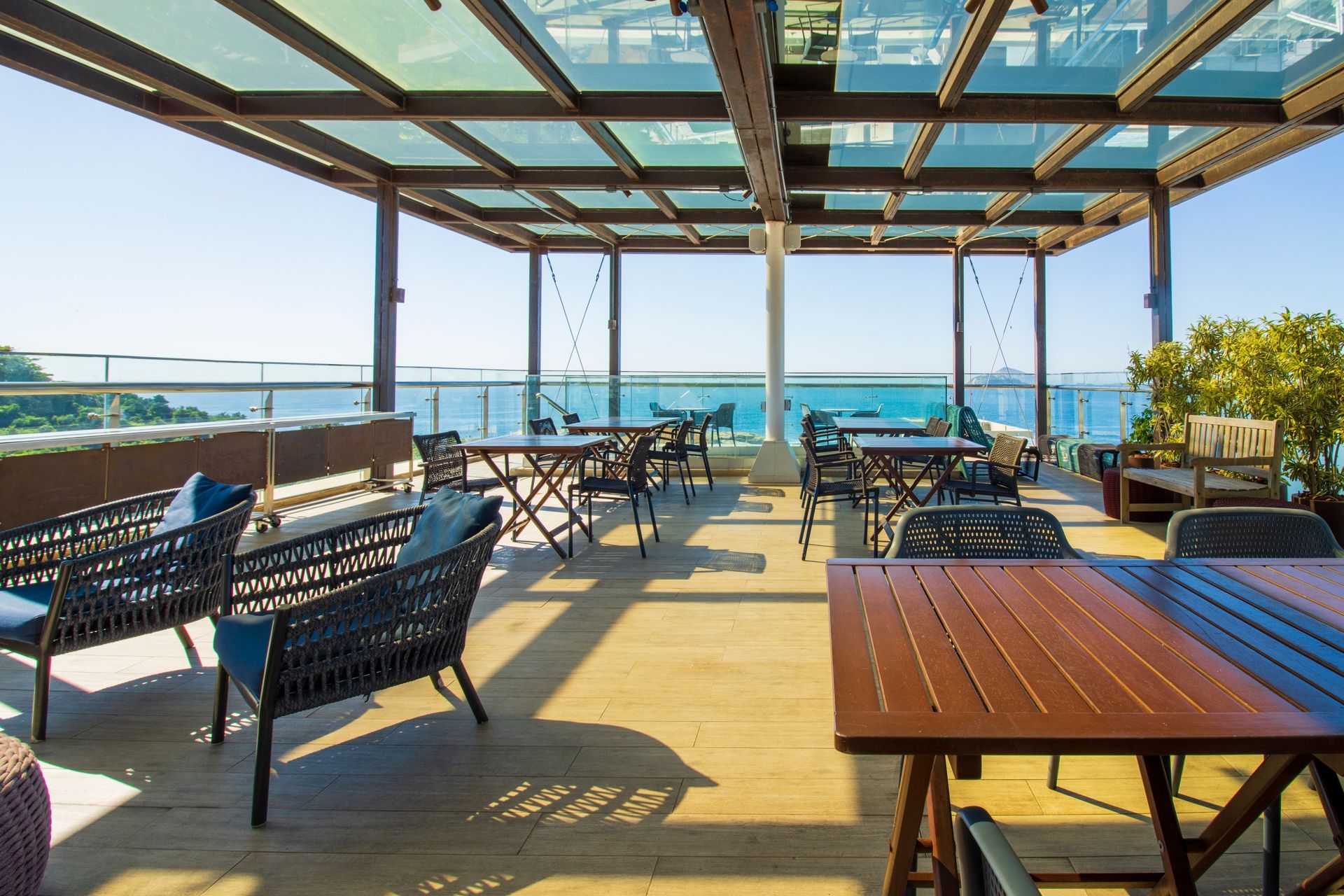 Terraço de restaurante à beira-mar com cadeiras pretas, mesas de madeira e vista para o oceano sob uma pérgola.