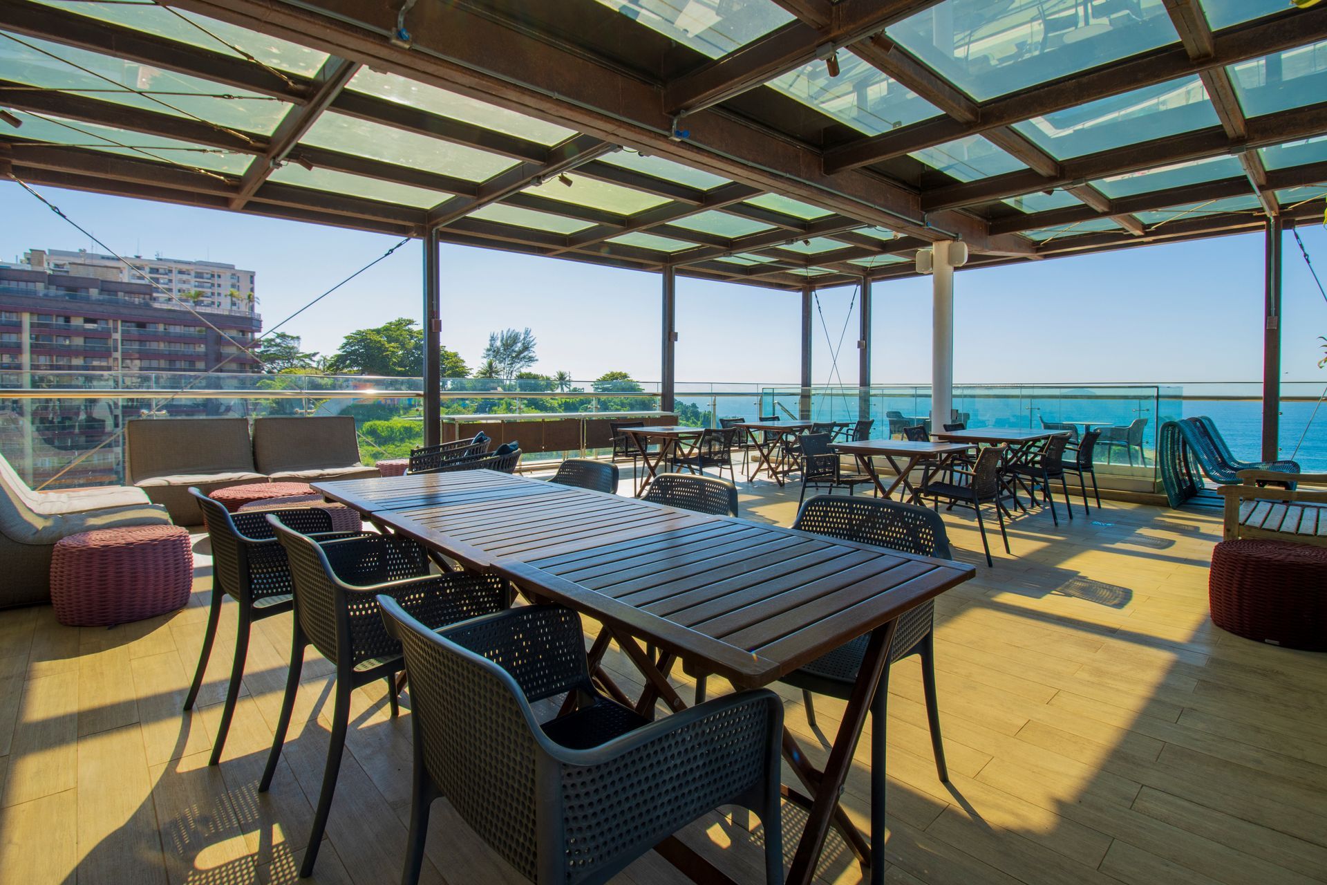 Café ao ar livre à beira-mar com mesas e cadeiras pretas sob uma pérgola de madeira, com vista para o oceano.