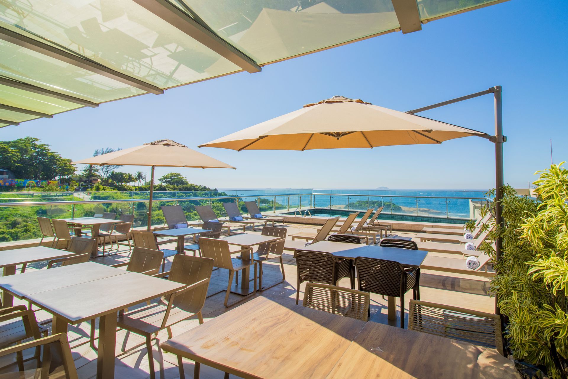 Terraço ensolarado de um café à beira-mar, com mesas de madeira, guarda-sóis e vista para o oceano.