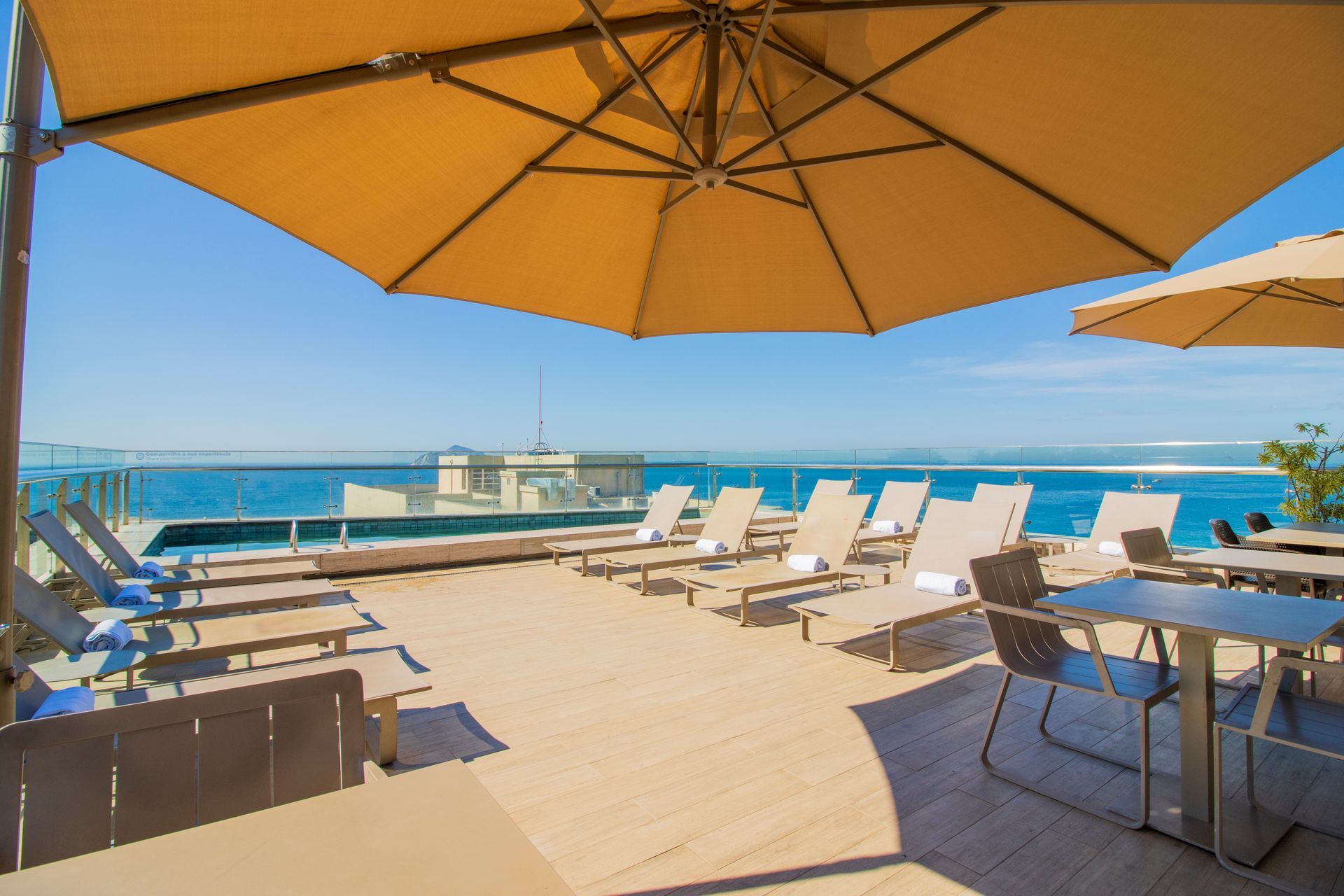 Terraço ensolarado à beira-mar com espreguiçadeiras brancas, mesas e grandes guarda-sóis com vista para o oceano.