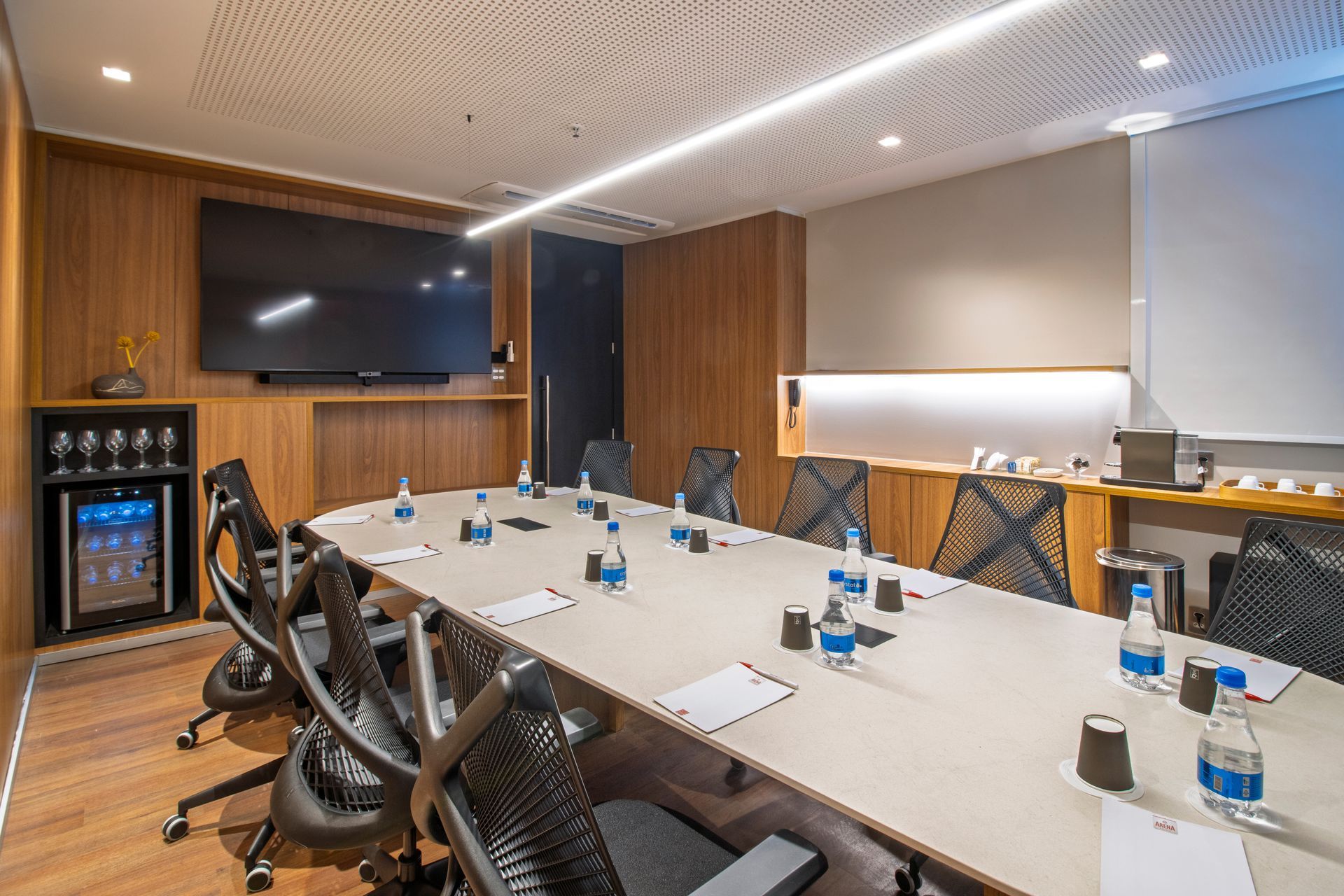 Sala de conferências moderna com mesa comprida, cadeiras giratórias, TV de parede e garrafas de água.