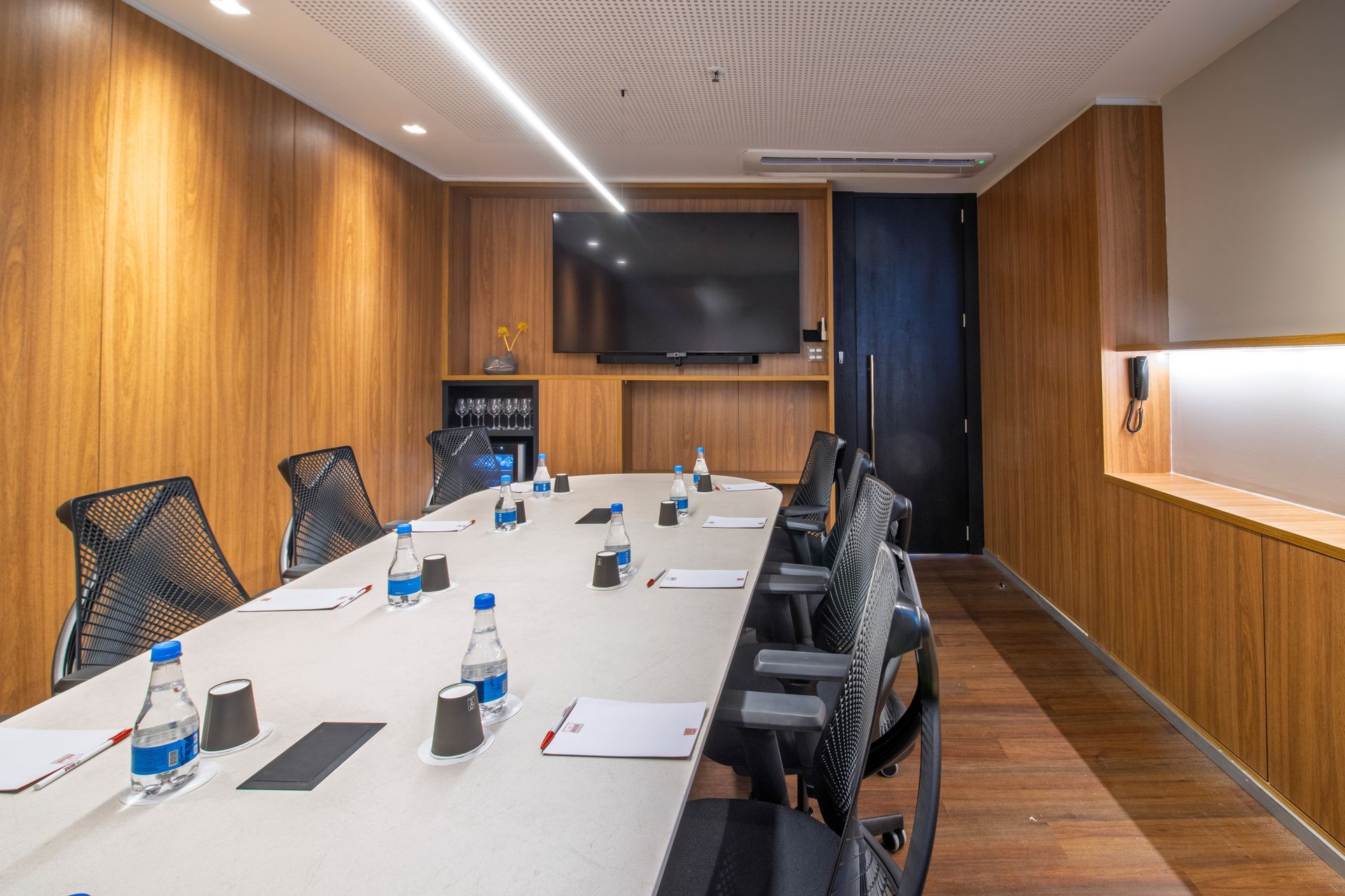 Sala de conferências moderna com mesa comprida, cadeiras, TV fixada na parede e paredes revestidas de madeira.