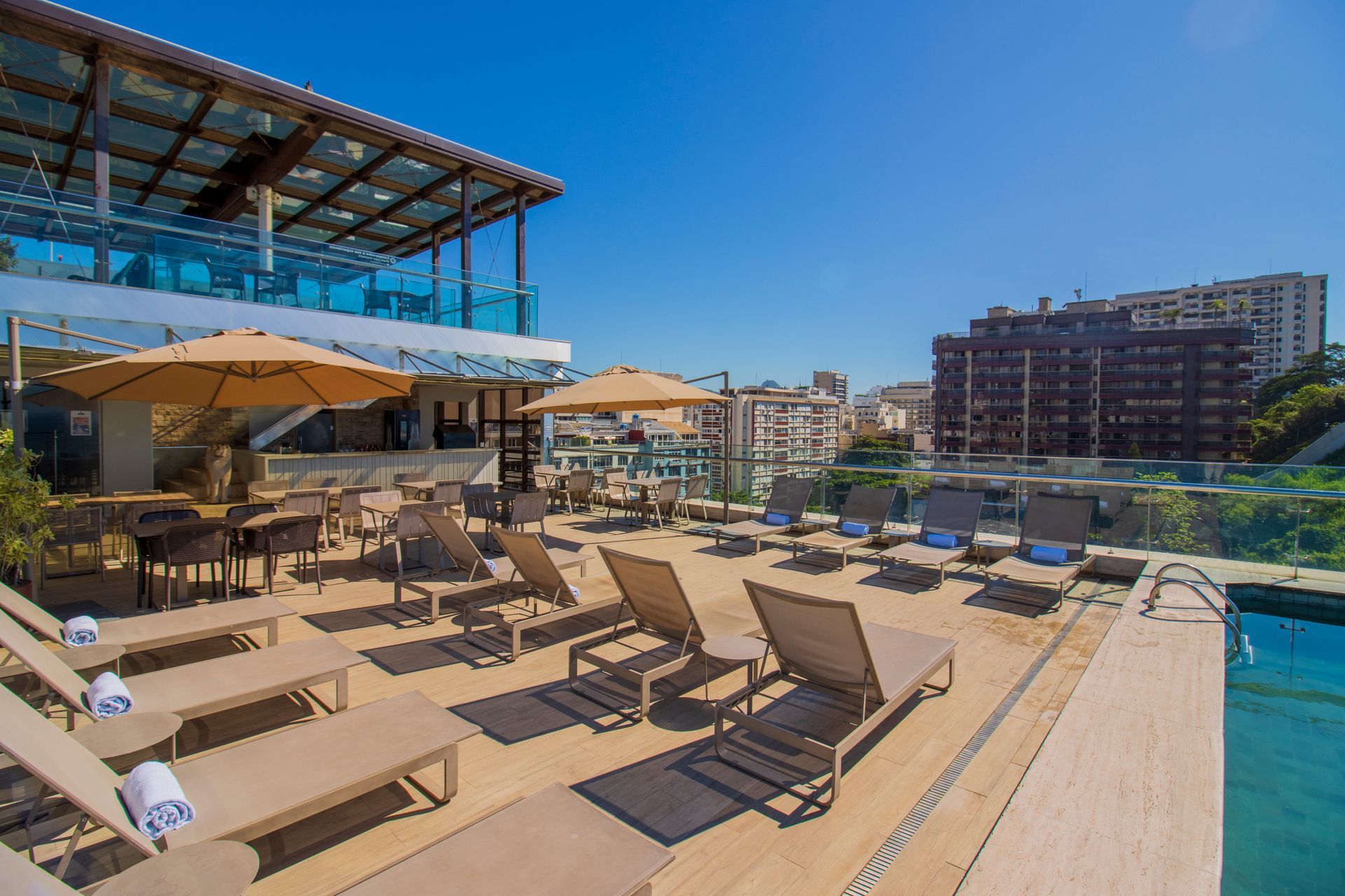 Terraço com piscina na cobertura, espreguiçadeiras, guarda-sóis e vista para os prédios da cidade sob um céu azul brilhante.