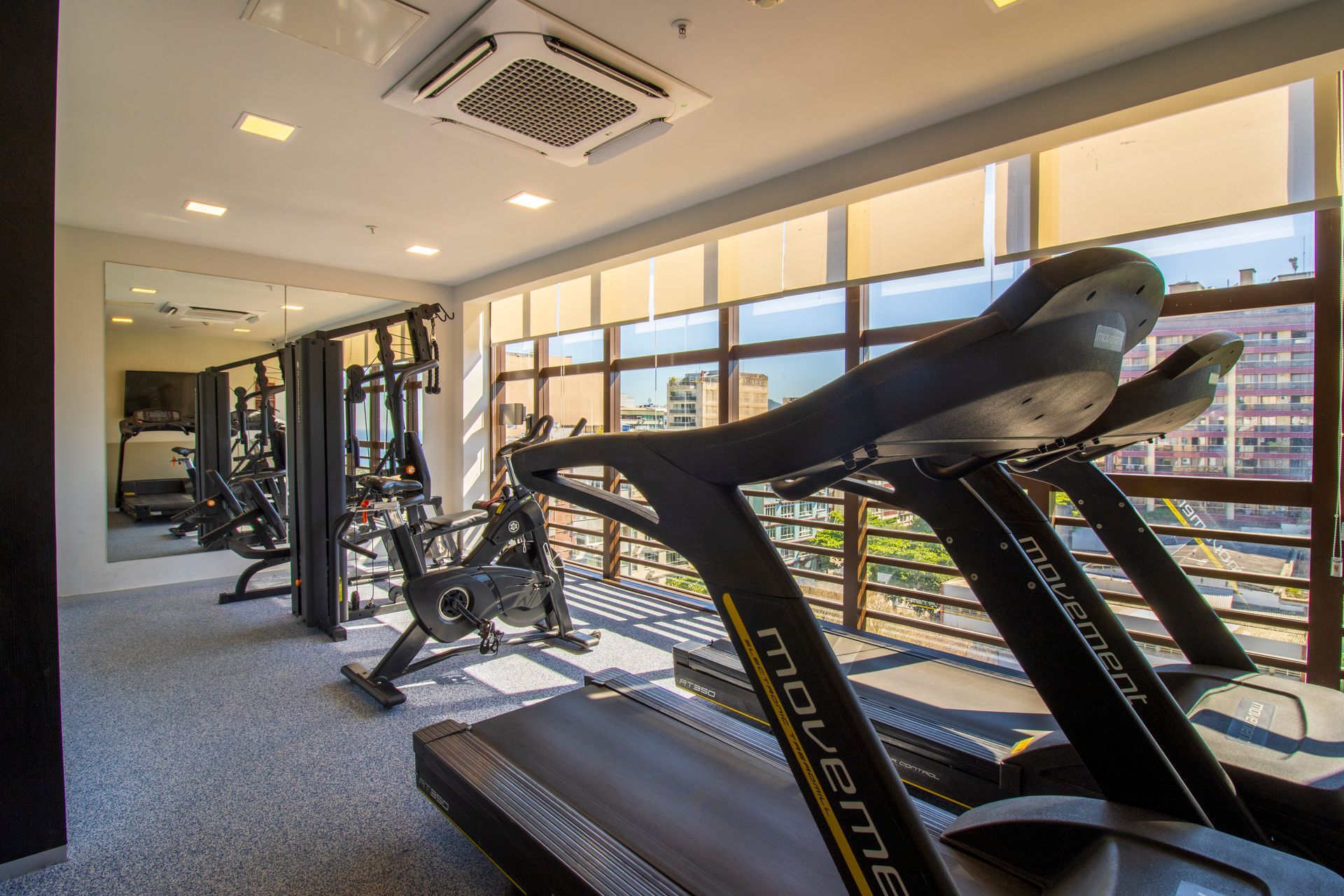 Academia moderna com esteiras e aparelhos de musculação, janelas amplas e vista para a cidade.