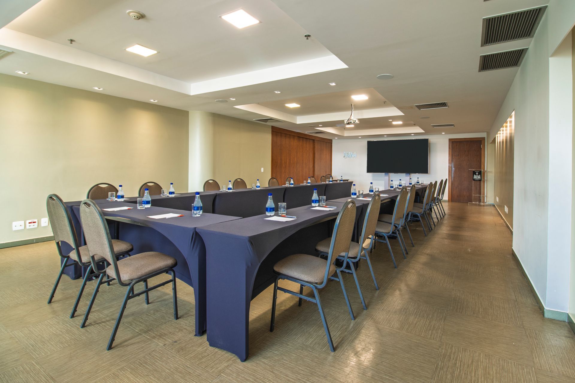 Sala de conferências com mesa em formato de U, toalhas de mesa azuis, cadeiras e tela fixada na parede, sob luzes brilhantes no teto.
