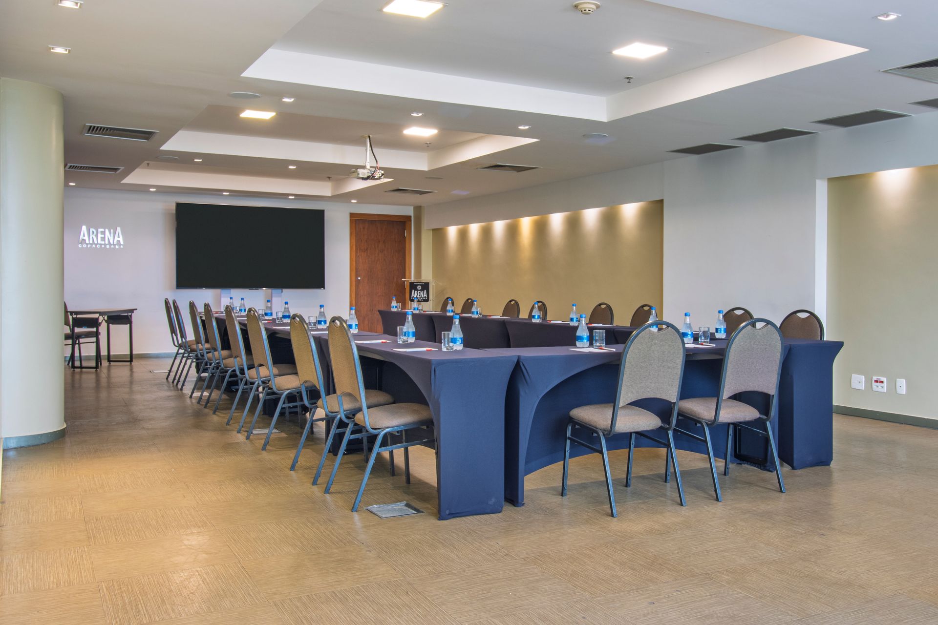 Sala de conferências com mesas compridas, cadeiras e uma tela fixada na parede em um ambiente de escritório moderno.