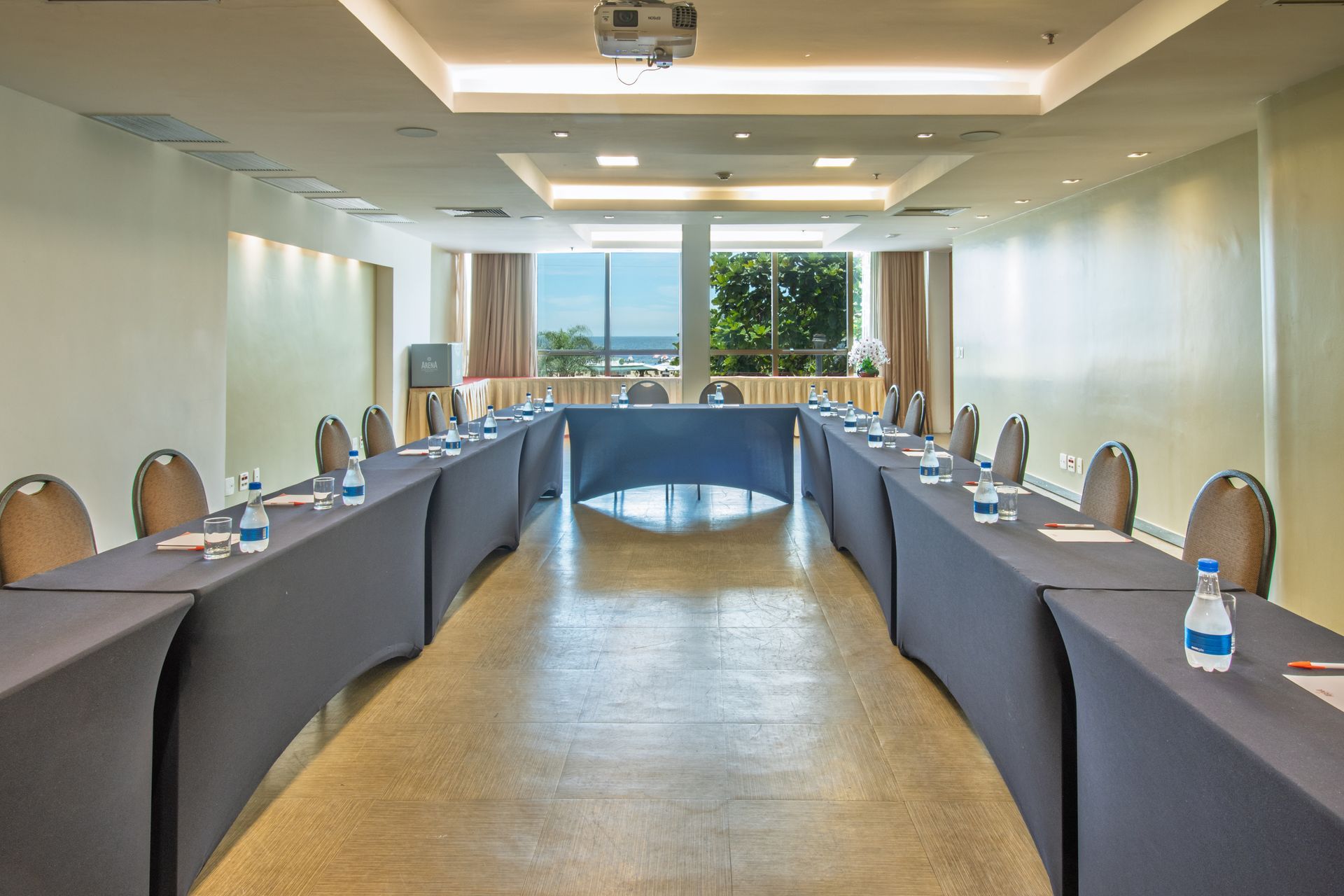 Sala de conferências com mesas em formato de U, garrafas de água e cadeiras voltadas para uma janela bem iluminada.
