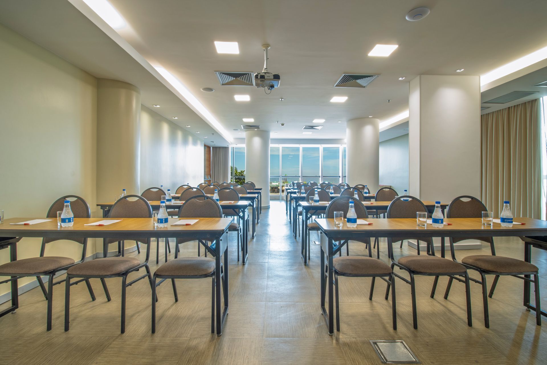Sala de conferências vazia com fileiras de mesas e cadeiras, luzes fortes e grandes janelas ao fundo.