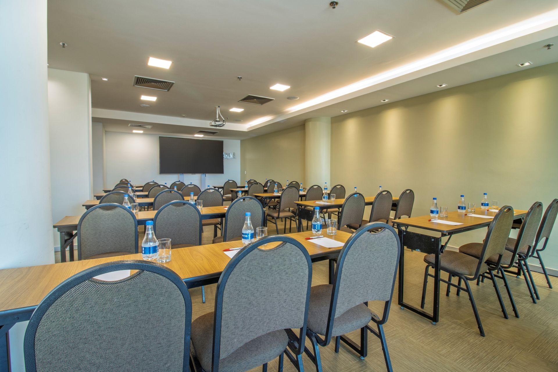 Sala de conferências vazia com fileiras de mesas e cadeiras, tela de projeção e iluminação forte no teto.