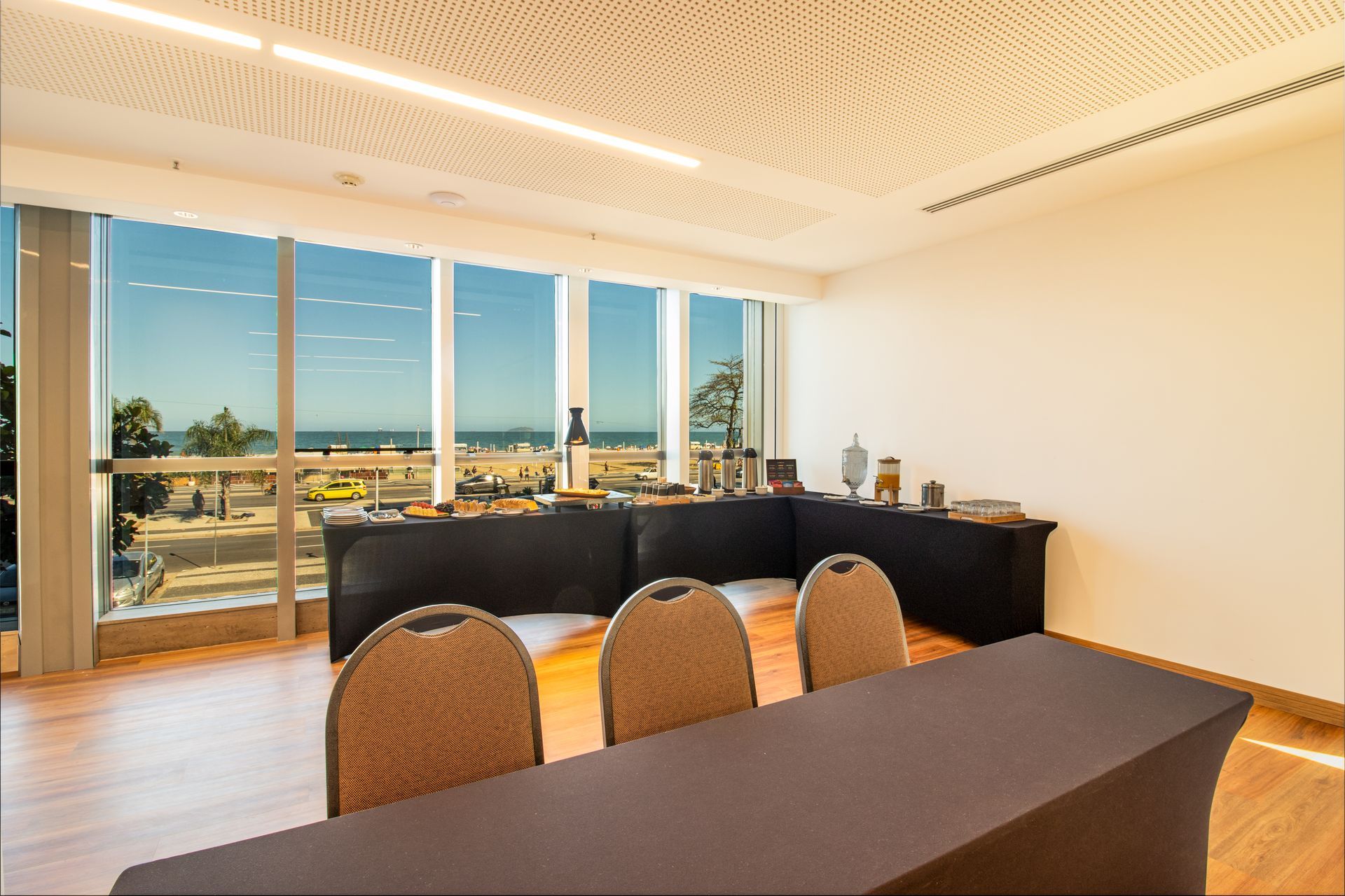 Sala de conferências iluminada com grandes janelas com vista para a cidade, mesa preta e cadeiras de vime.