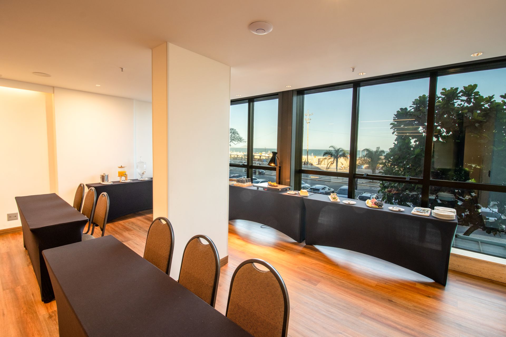 Sala de jantar moderna com longas mesas pretas, cadeiras de vime e grandes janelas com vista para o horizonte da cidade.