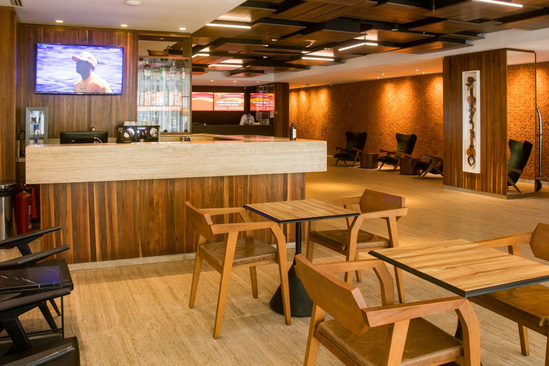 Área de estar vazia de um café moderno com mesas de madeira, balcão branco e televisões fixadas na parede.