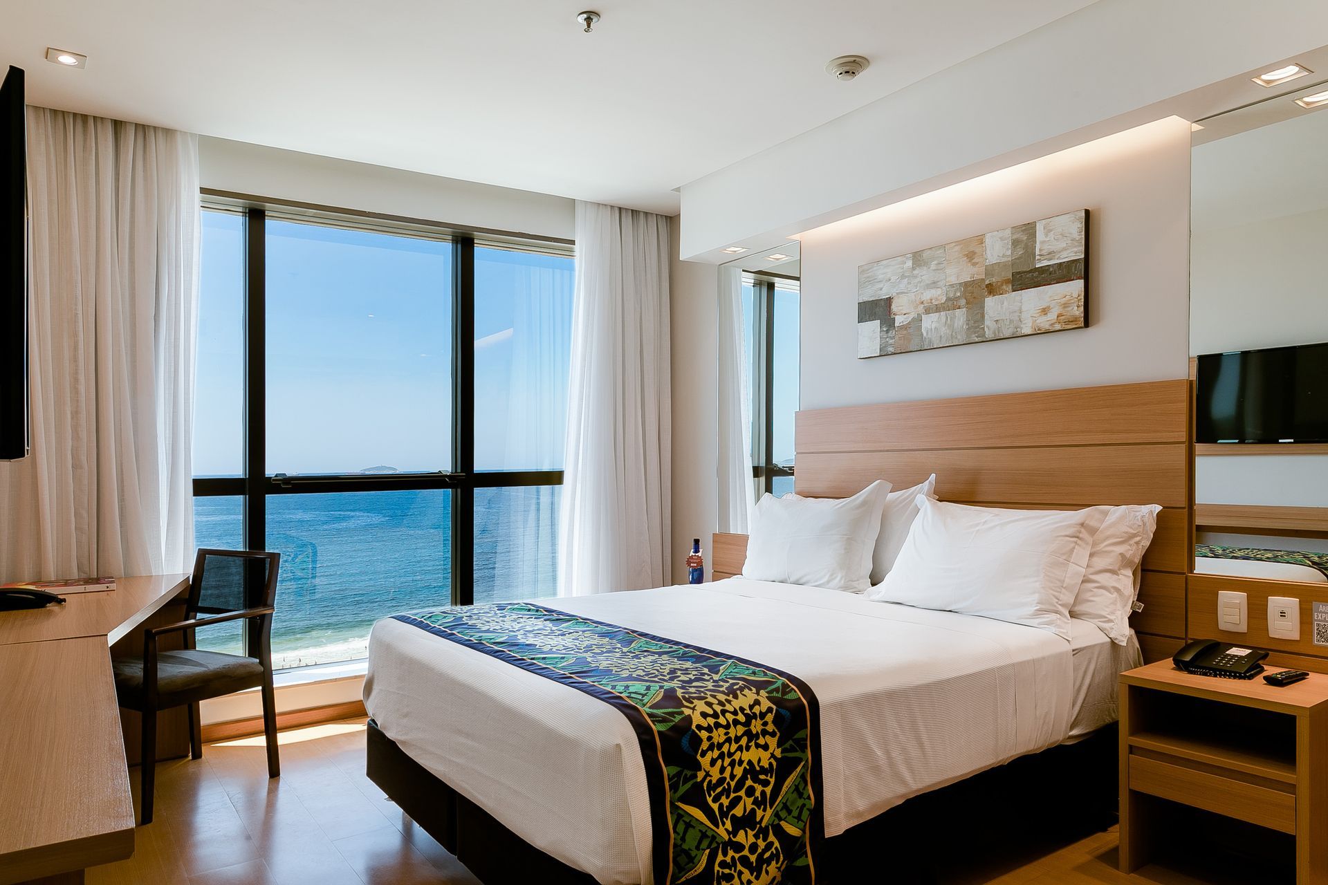 Quarto de hotel moderno com cama king-size, vista para o mar e janelas panorâmicas.