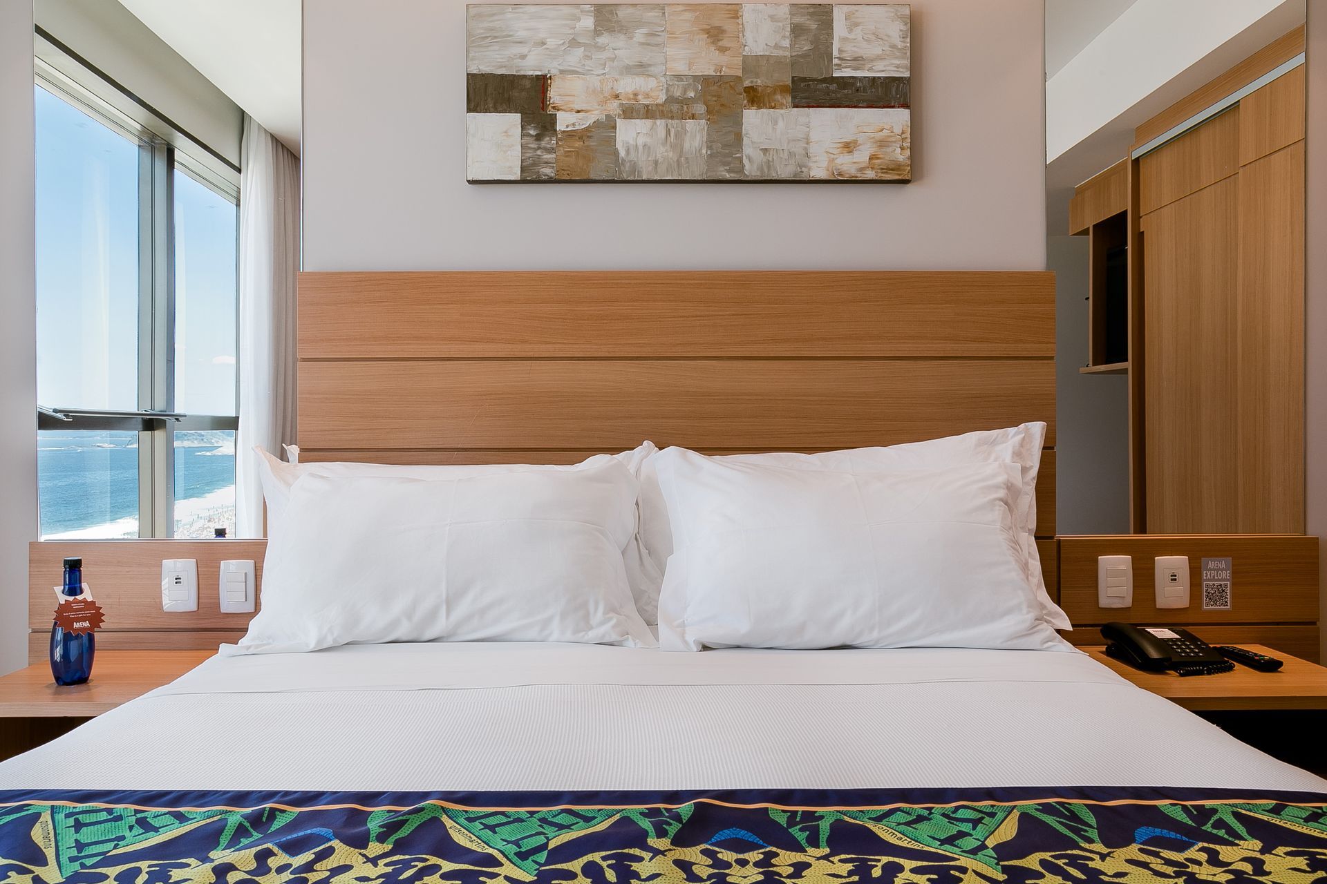 Quarto de hotel moderno com cama bem arrumada, vista para o mar e cabeceira de madeira.