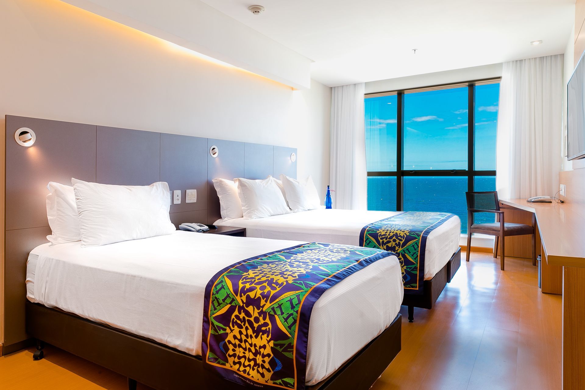 Quarto de hotel moderno com duas camas brancas, tapetes coloridos e uma janela com vista para o mar.