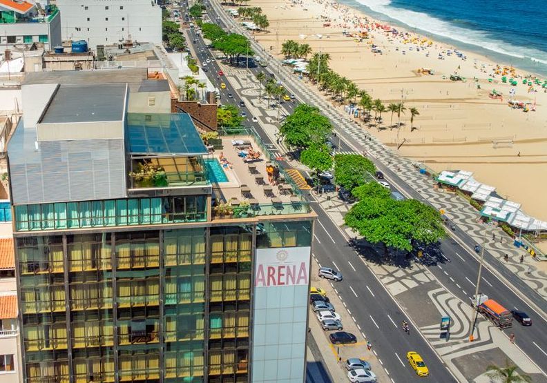 Vista aérea de uma rua litorânea com prédios altos, carros estacionados e uma praia de areia com guarda-sóis.