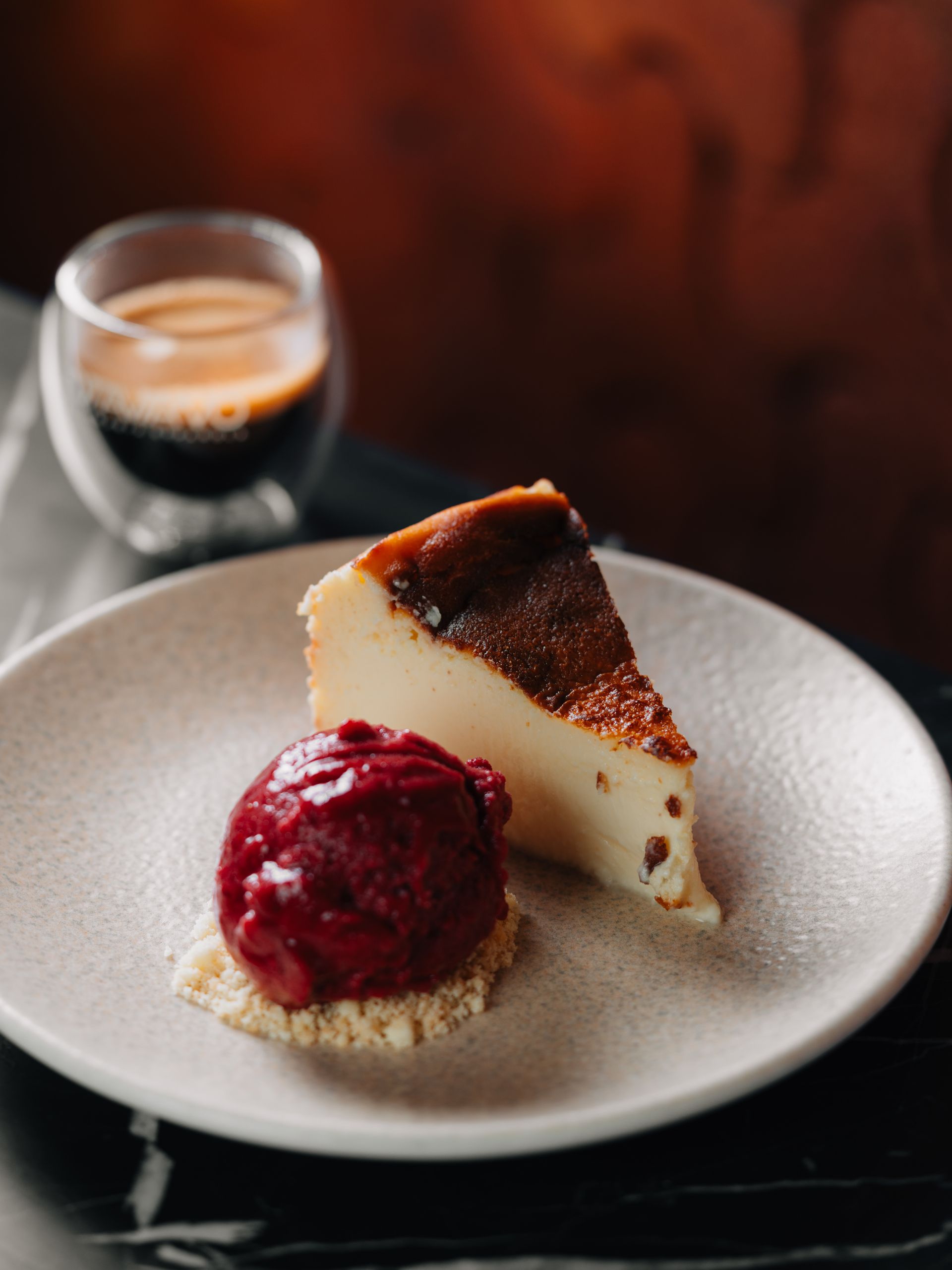 Fatia de cheesecake com sorvete de frutas vermelhas em um prato bege sobre uma mesa de madeira.