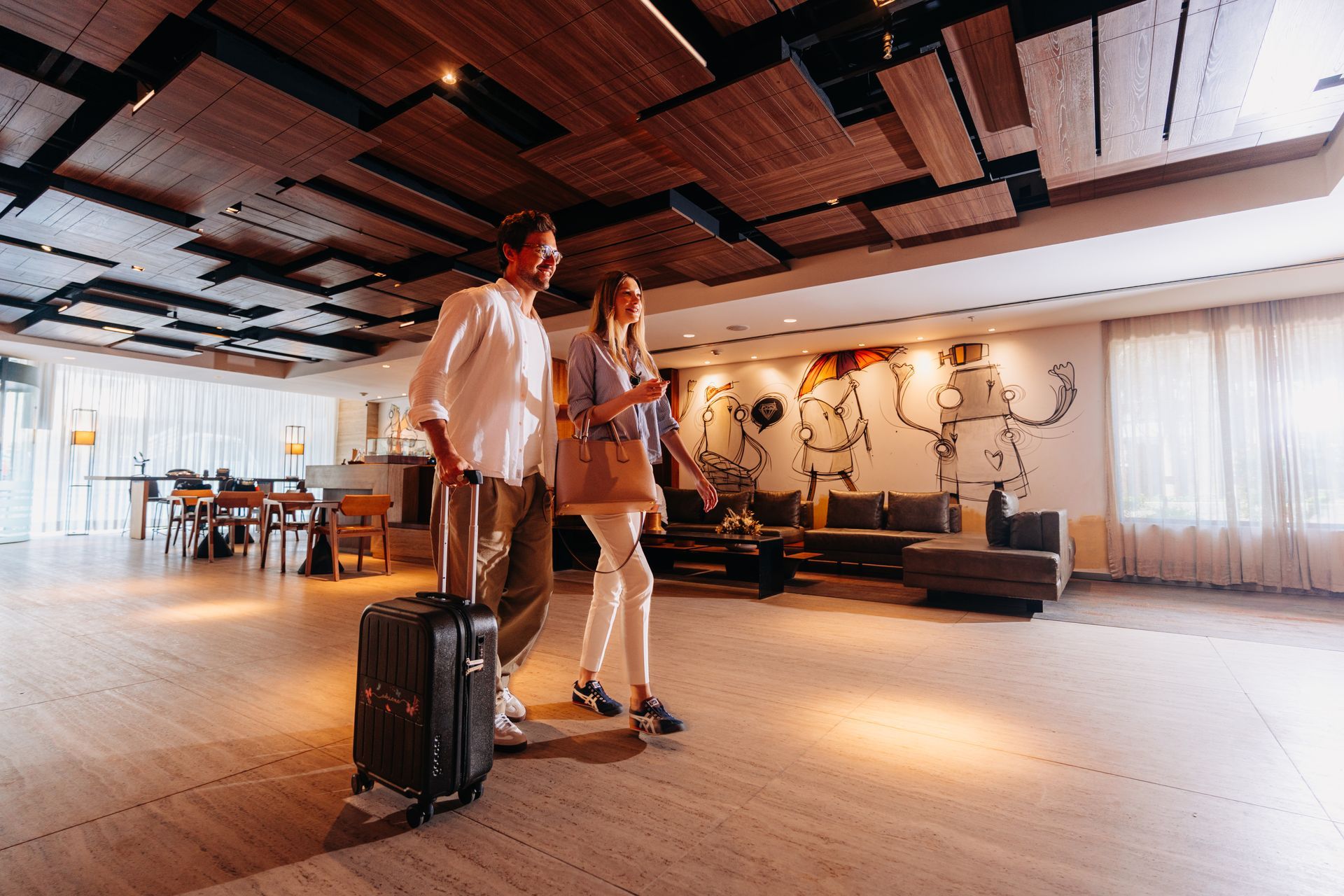 Dois viajantes com bagagem em um lounge moderno e iluminado com teto de madeira aparente.