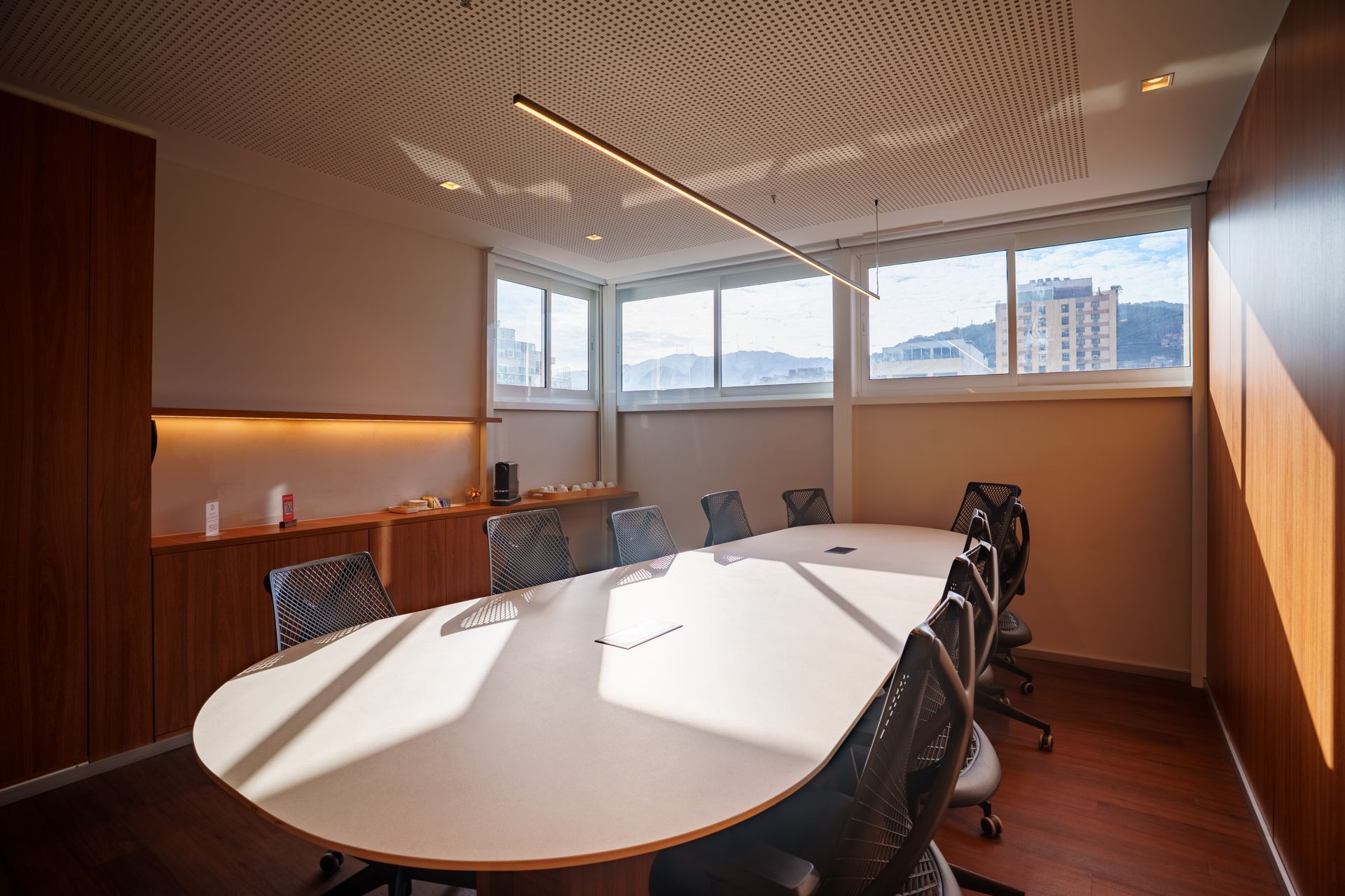Sala de conferências moderna com mesa oval, cadeiras pretas, painéis de madeira e grandes janelas com vista para a cidade.
