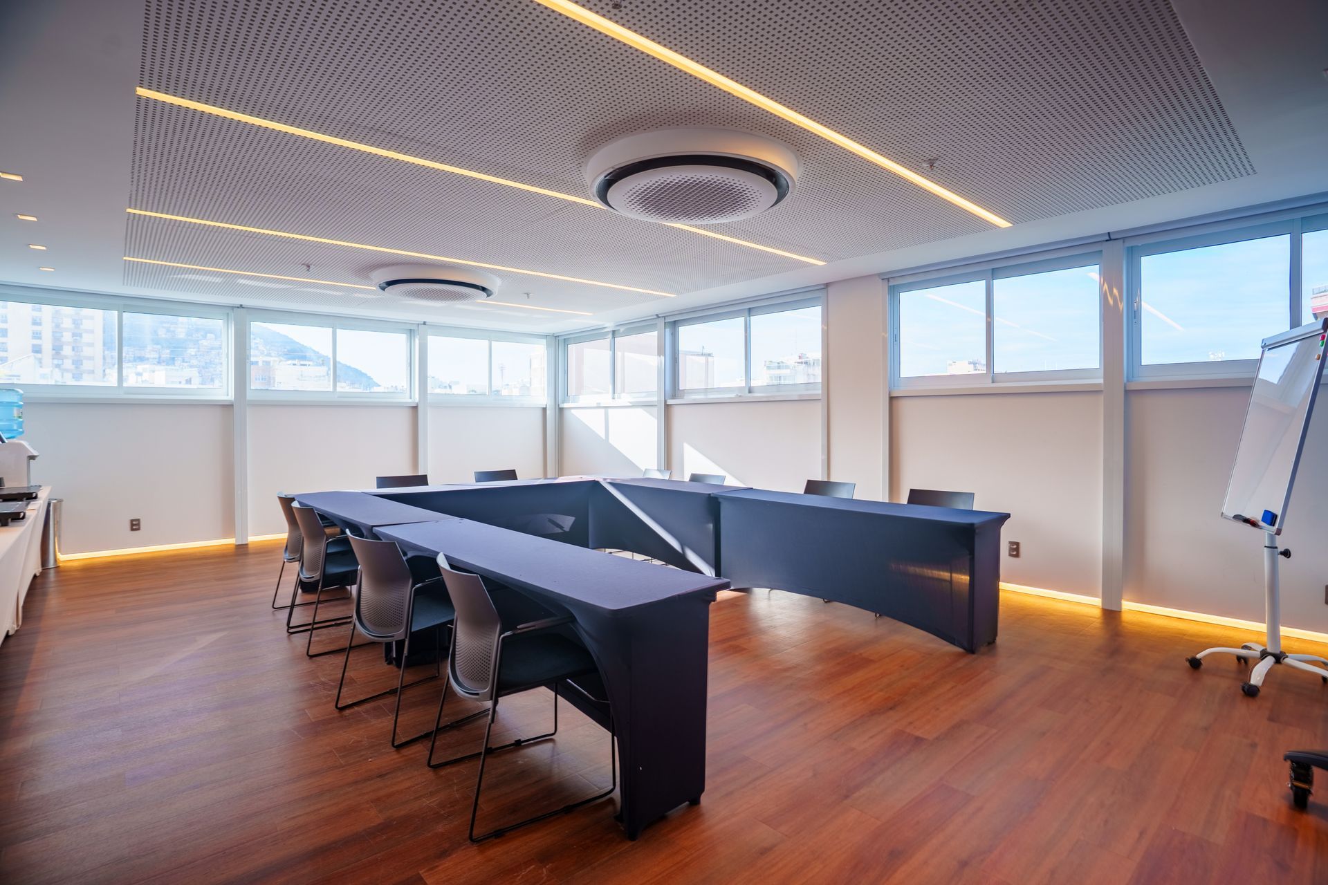 Sala de conferências moderna e vazia, com mesas e cadeiras pretas, piso de madeira e janelas amplas.