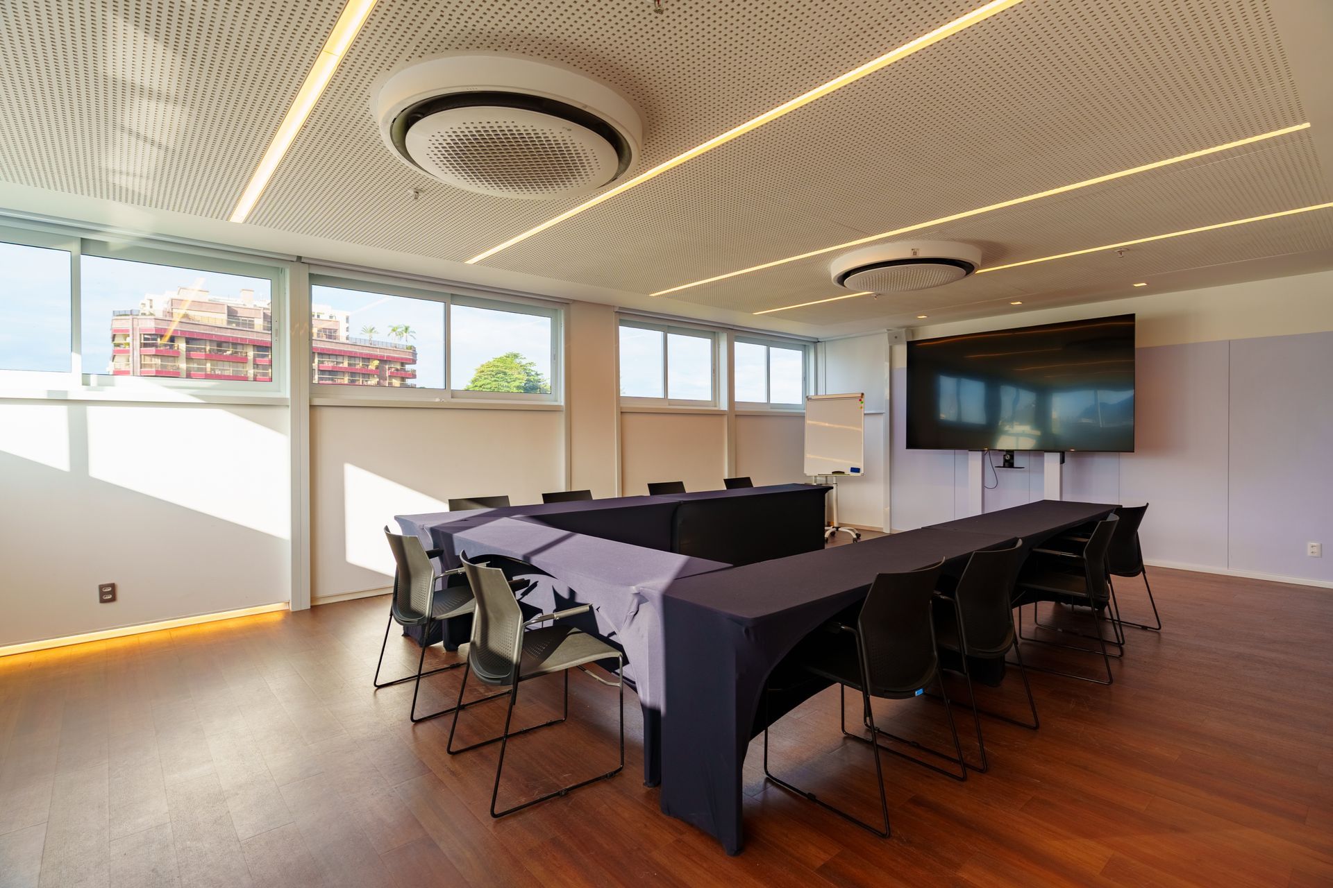 Sala de conferências moderna com mesa em formato de U, cadeiras pretas, TV fixada na parede e janelas amplas.