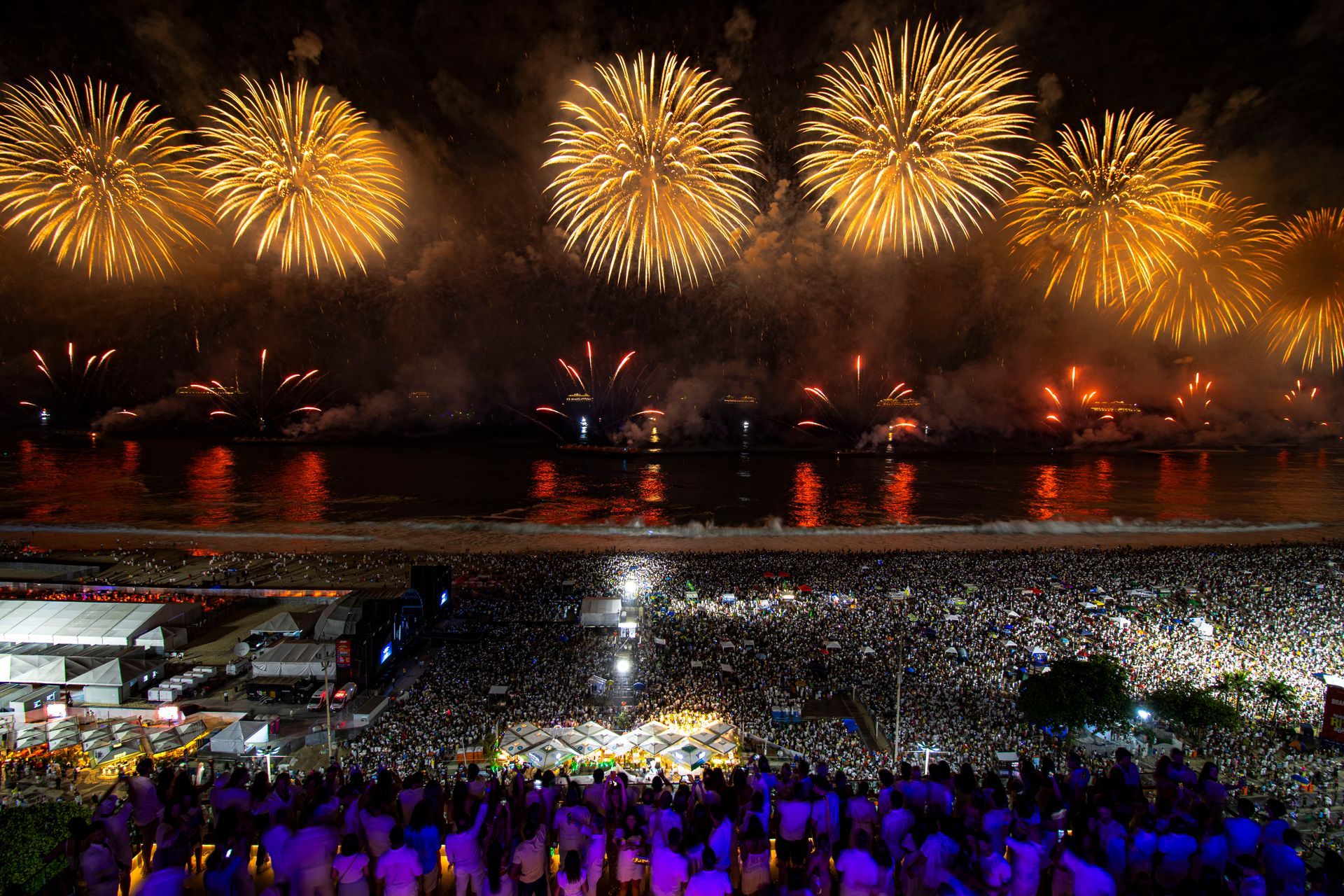 Fogos de artifício sobre um estádio lotado, com explosões douradas brilhantes e uma multidão iluminada de roxo abaixo.
