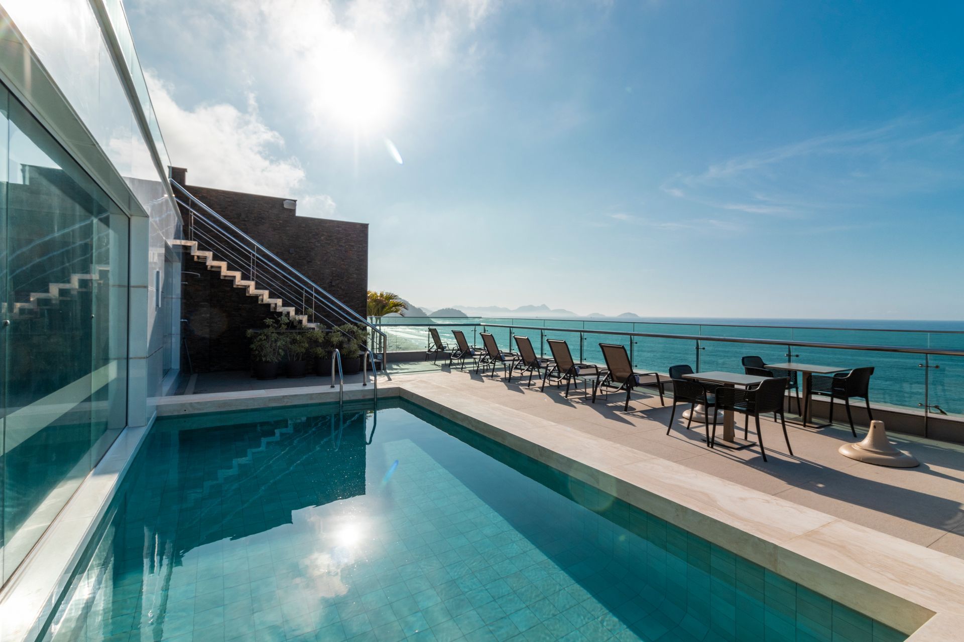 Terraço luxuoso à beira da piscina com vista para o oceano, com espreguiçadeiras e mesas sob um céu ensolarado.