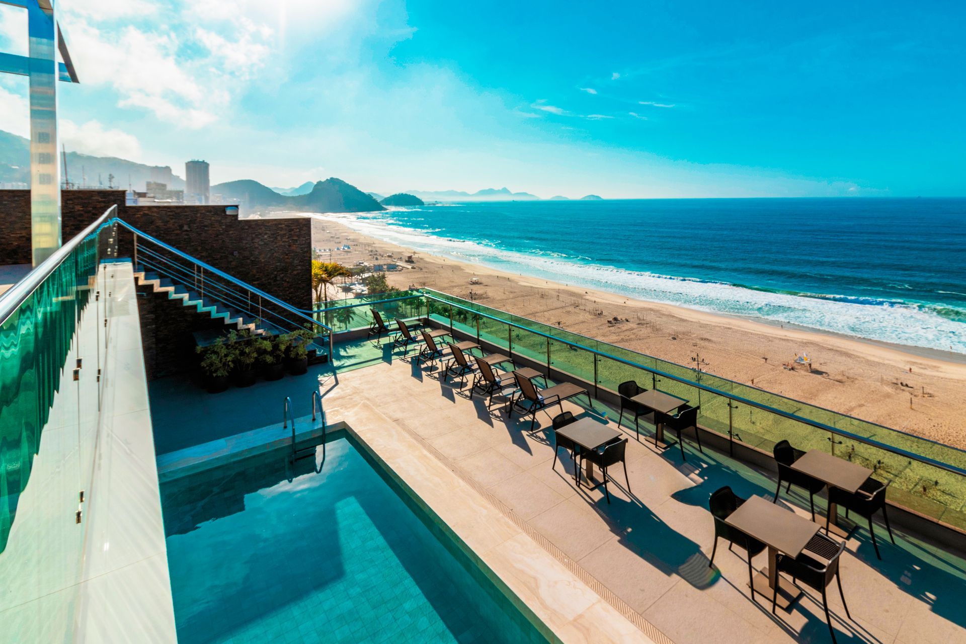 Terraço de resort à beira-mar com piscina, espreguiçadeiras e praia de areia sob um céu azul brilhante.
