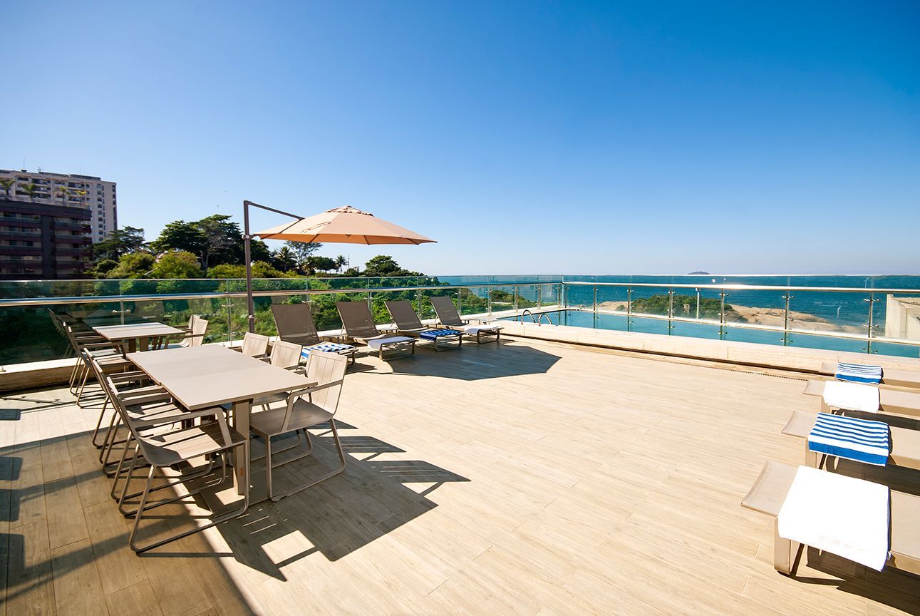 Terraço ensolarado à beira-mar com espreguiçadeiras, guarda-sol e vista para o oceano.