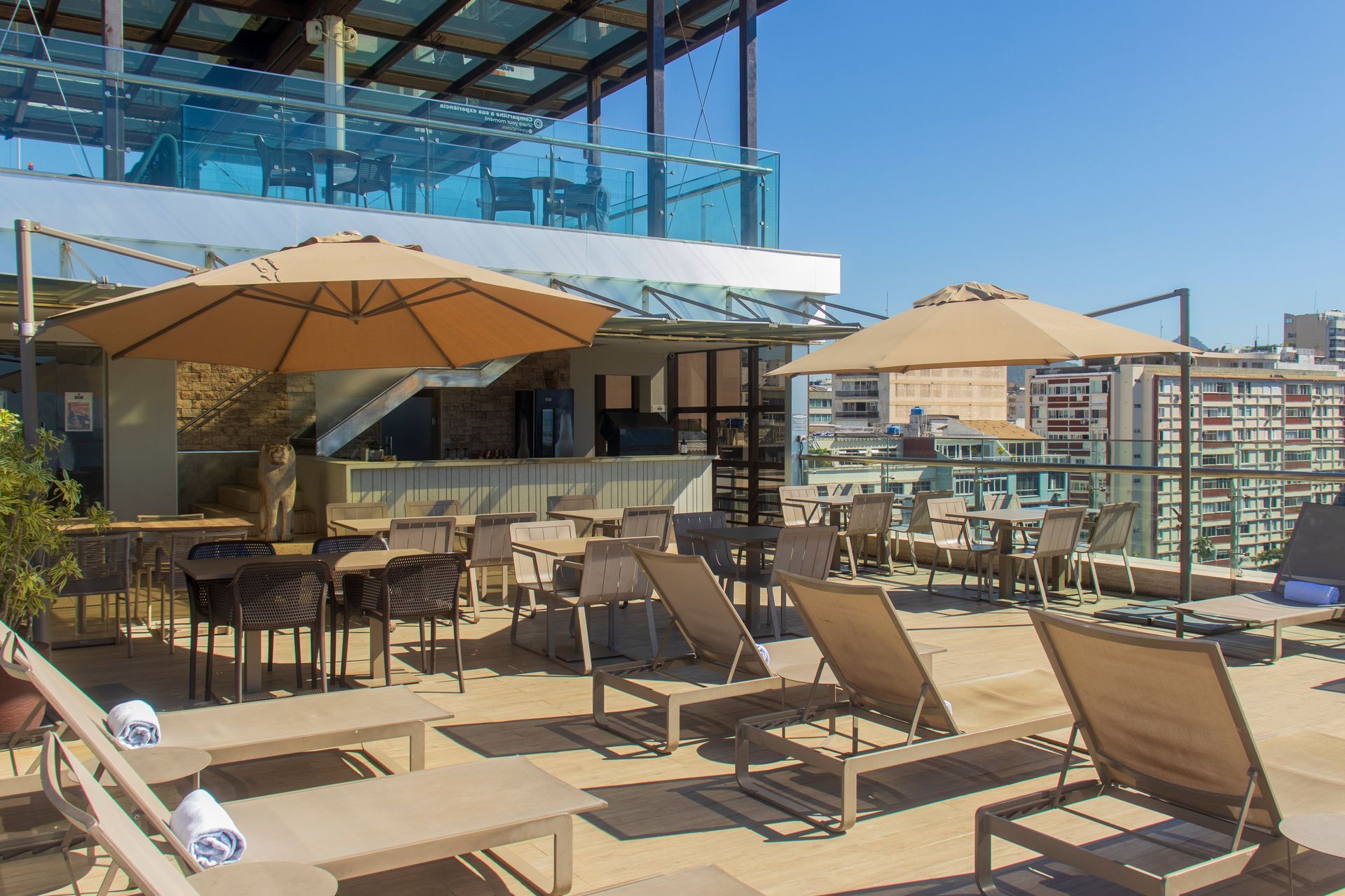 Terraço na cobertura com espreguiçadeiras, mesas, guarda-sóis e vista para a cidade sob um toldo de vidro.