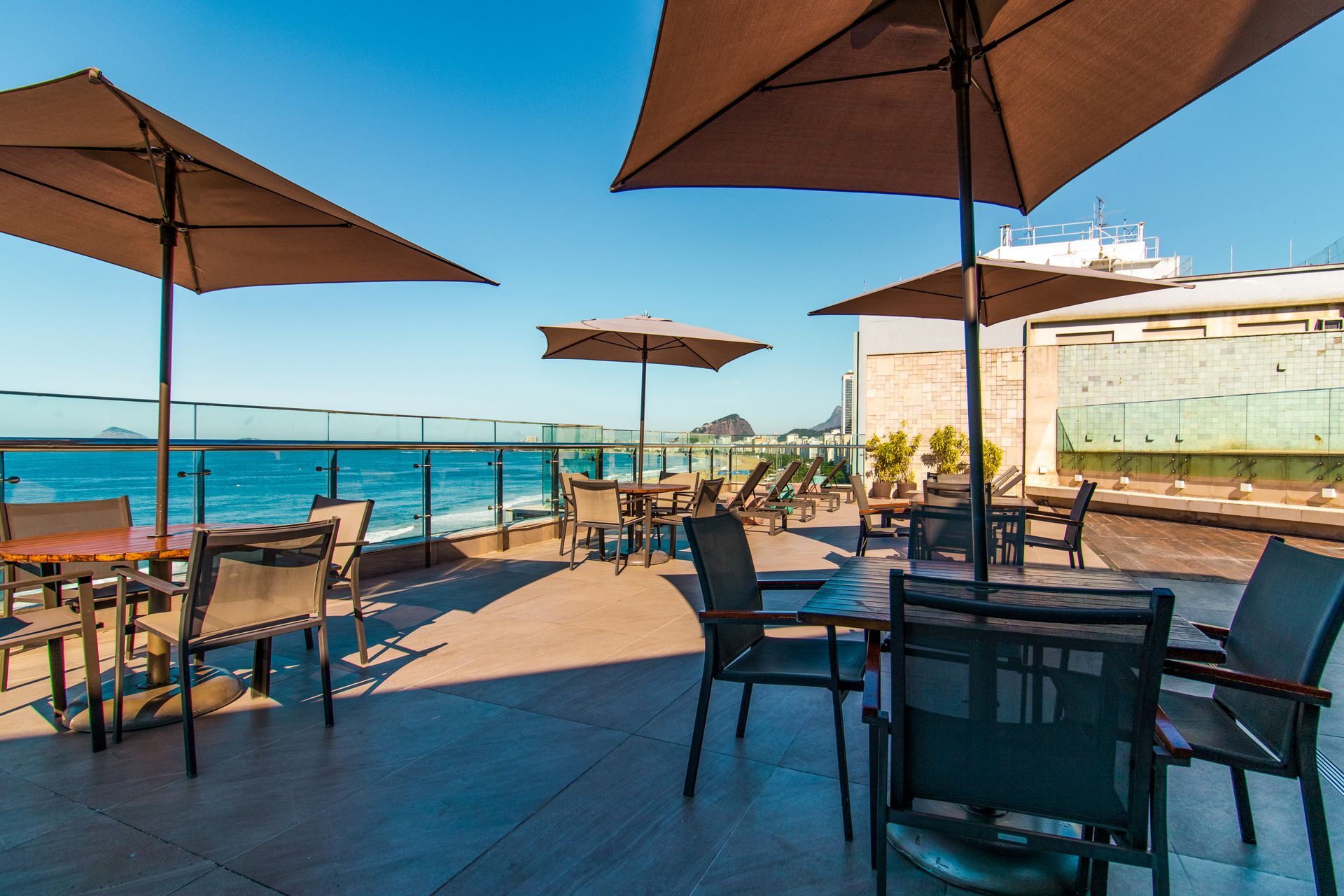 Terraço de café ao ar livre à beira-mar com mesas, guarda-sóis e vista para o oceano sob um céu azul brilhante.