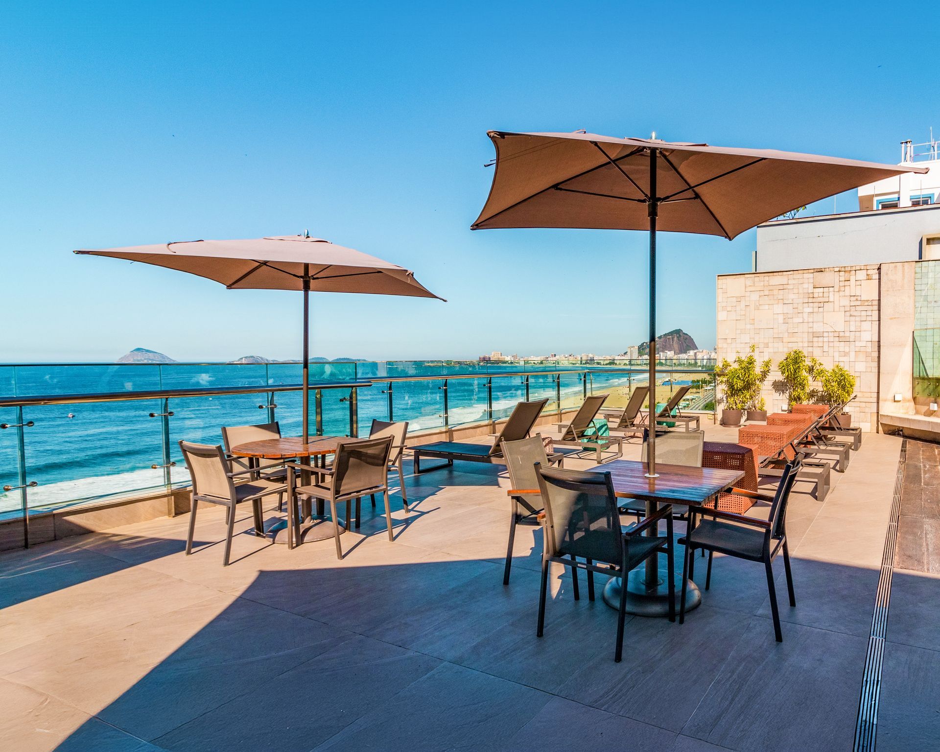 Terraço de café à beira-mar com mesas, guarda-sóis e vista para o oceano sob um céu azul límpido.